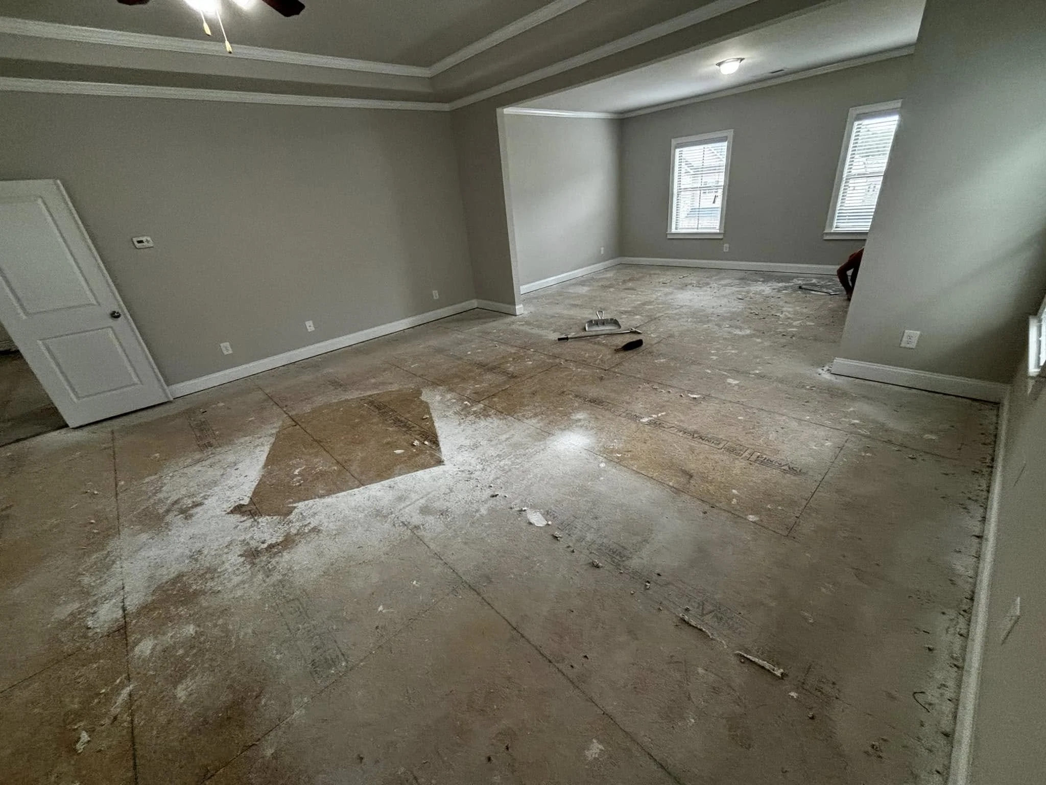 Empty living room under renovation with exposed subflooring, light gray walls, three windows with blinds, and a ceiling with a fan.