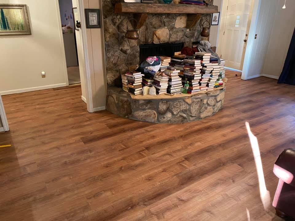 Living room with hardwood flooring, a stone fireplace with a wooden mantel, and stacks of books on the hearth.