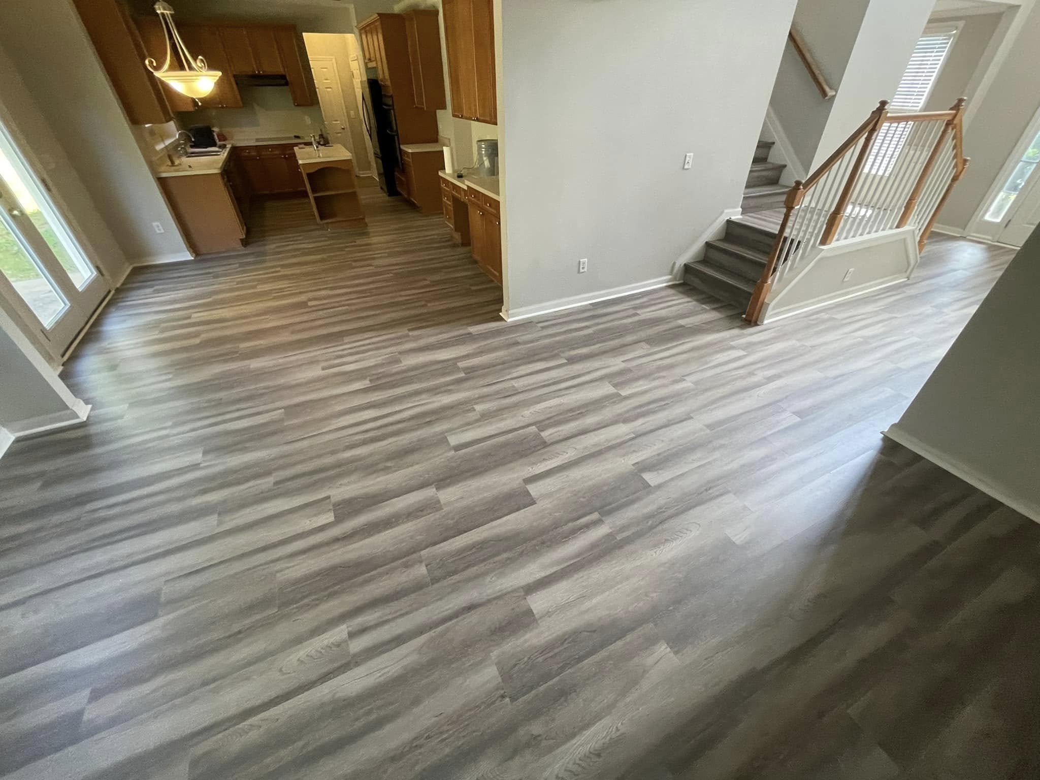 A spacious living area with light-colored walls, wood flooring, and a staircase with wooden railings. The kitchen is visible with wooden cabinets, a white countertop, and black appliances. There is a glass door leading outside and a large window allo