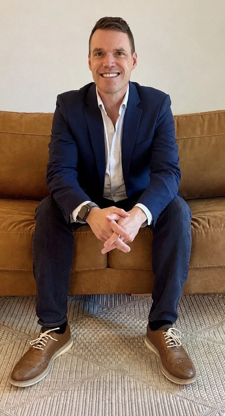 Man in a navy blazer and white shirt sitting on a brown couch, smiling with hands clasped, wearing brown dress shoes and casual pants.