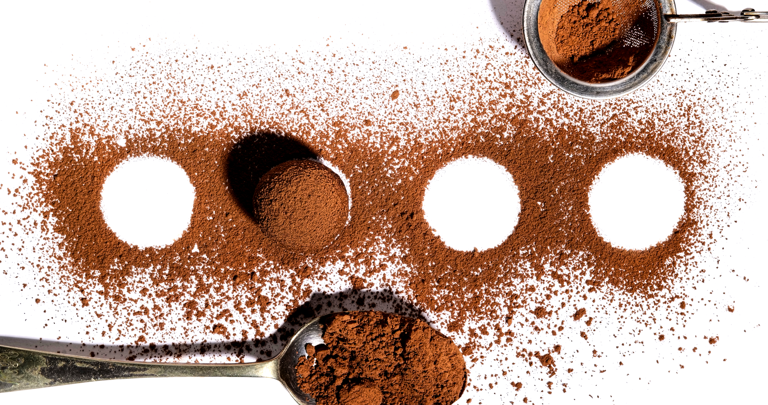 Spoons filled with cocoa powder surrounded by scattered cocoa powder on a white surface.