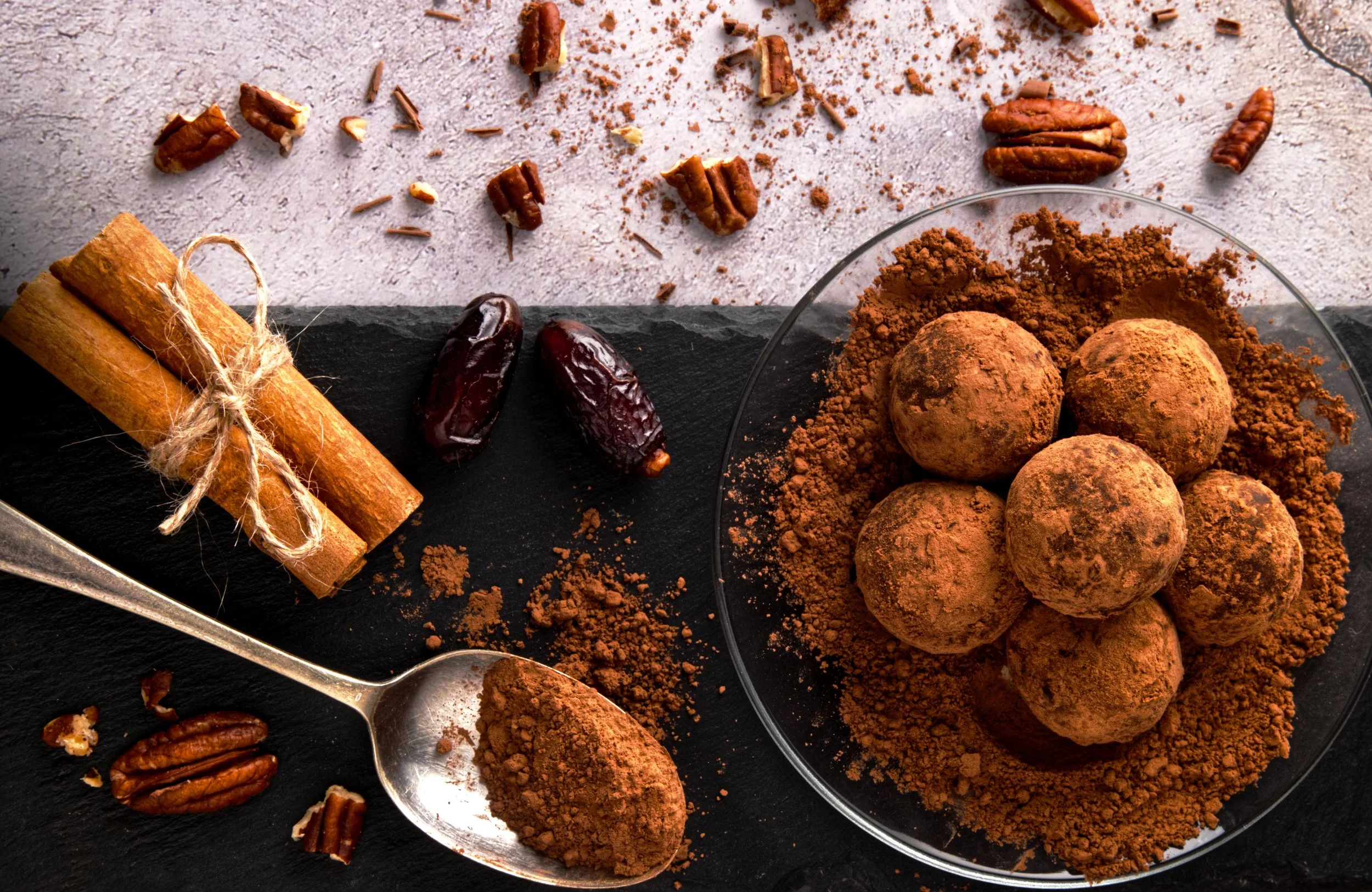 Chocolate truffles dusted with cocoa powder, walnuts, dates, and cinnamon sticks on a black slate and glass plates.
