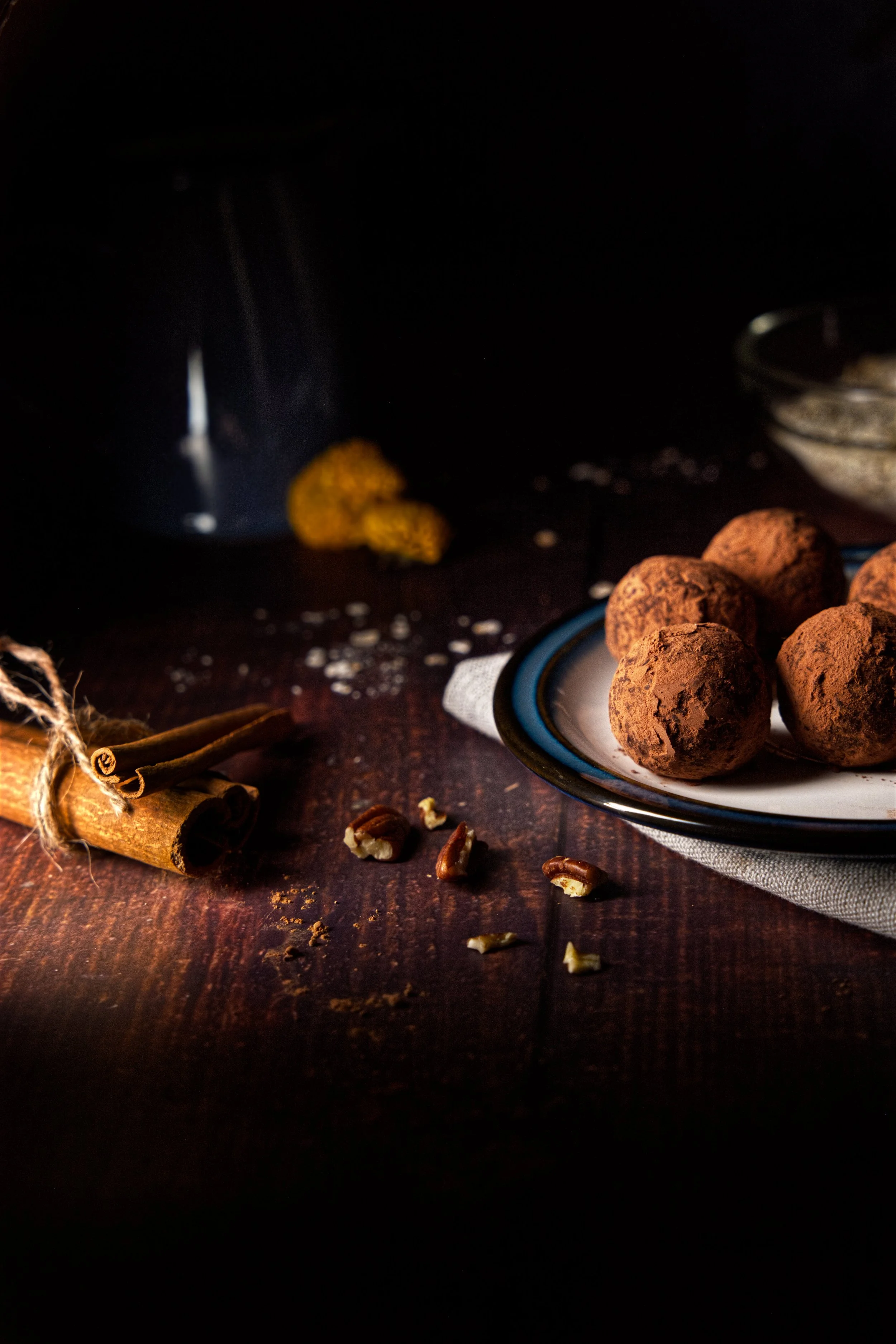 Chocolate truffles on a white plate with blue rim, cinnamon sticks, walnuts, and a jar of chopped nuts, dark background.