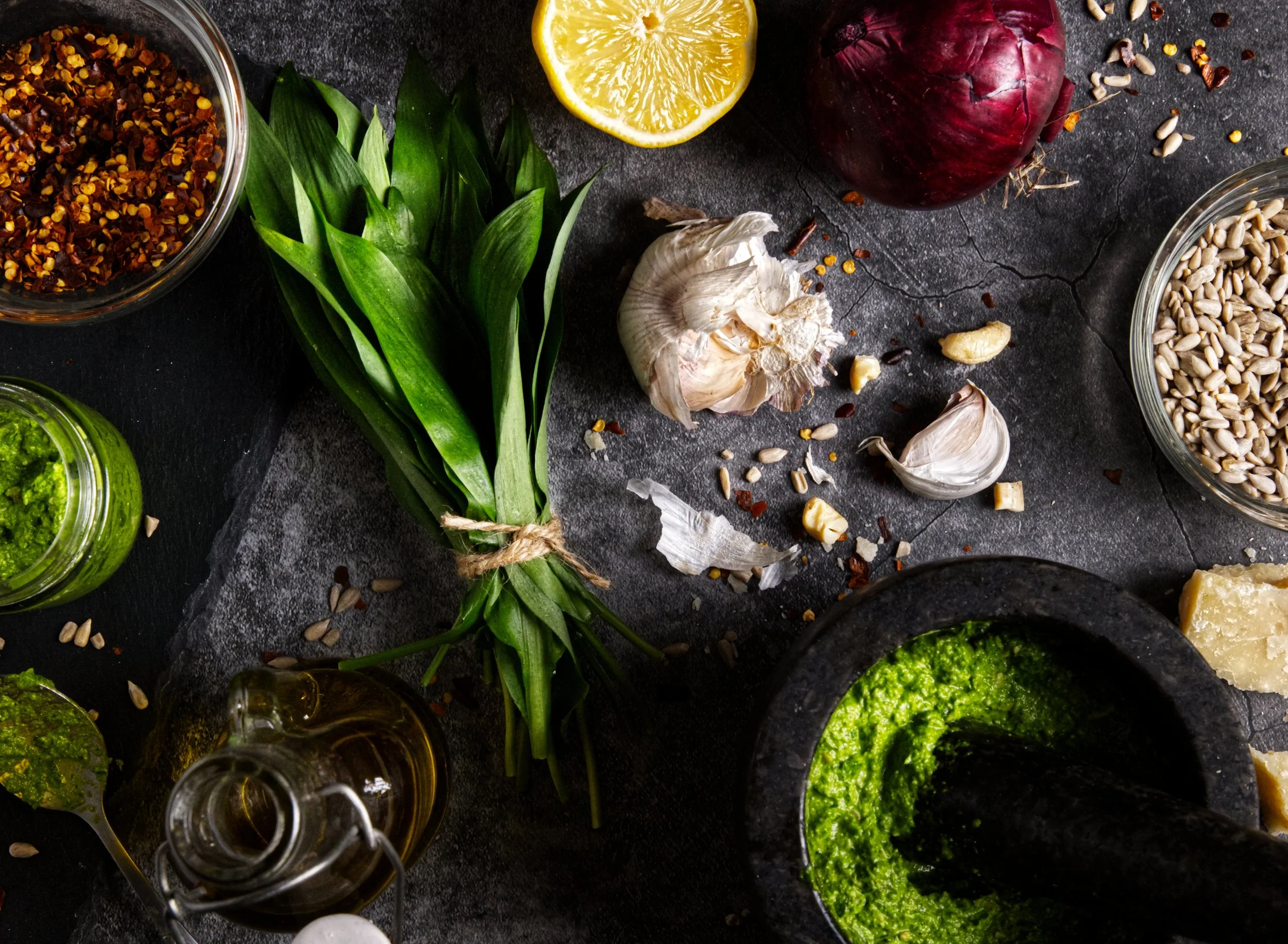 Fresh ingredients including lemon, garlic, red onion, basil, sunflower seeds, chili flakes, and pesto spread on a dark surface, with small jars of pesto and oil nearby.