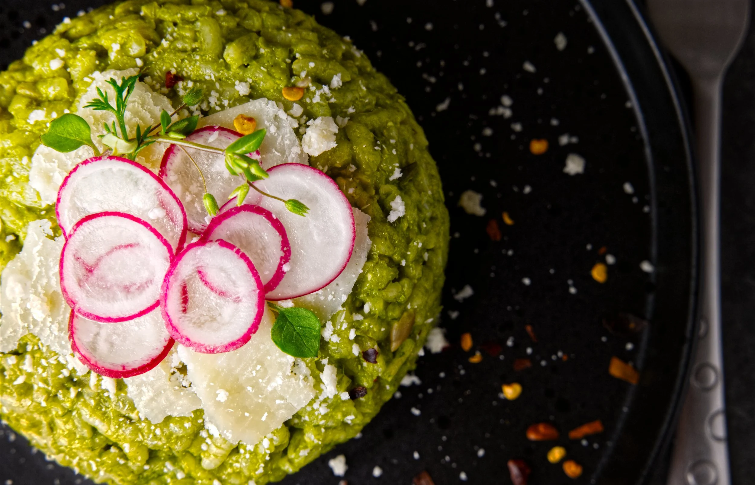 Close-up of a green dish garnished with sliced radishes, creamy white sauce, and herbs, served on a black plate.