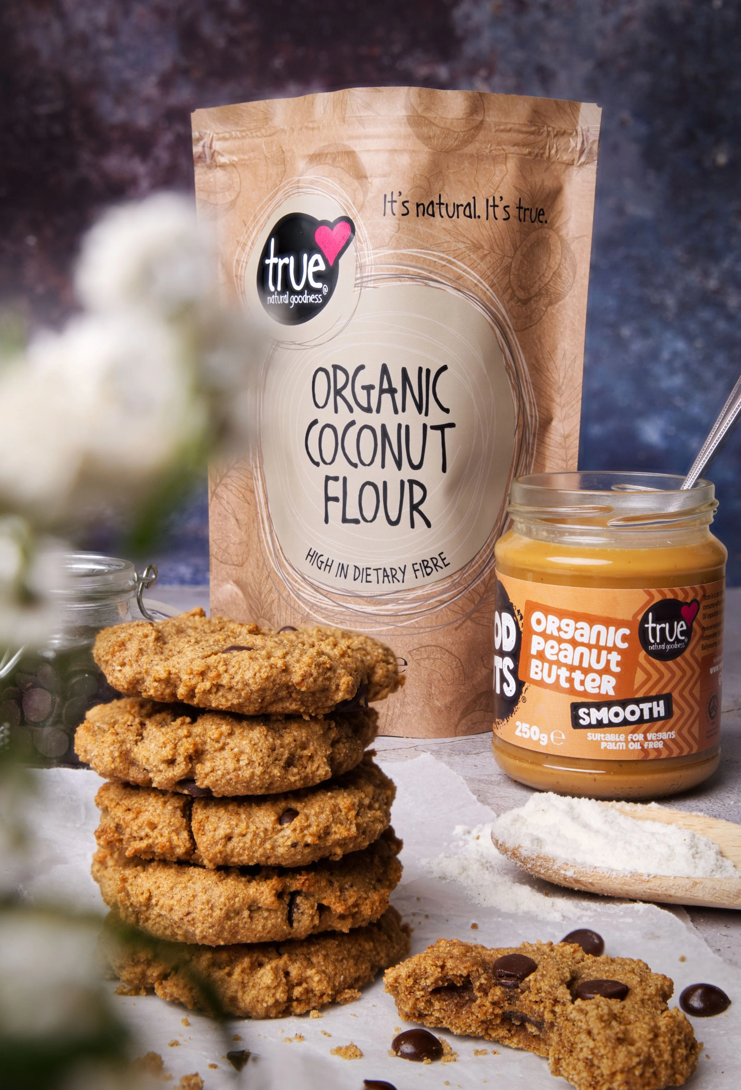 Stack of homemade chocolate chip cookies, jar of organic peanut butter, and a bag of organic coconut flour with the label 'True', placed on a white surface with scattered chocolate chips.