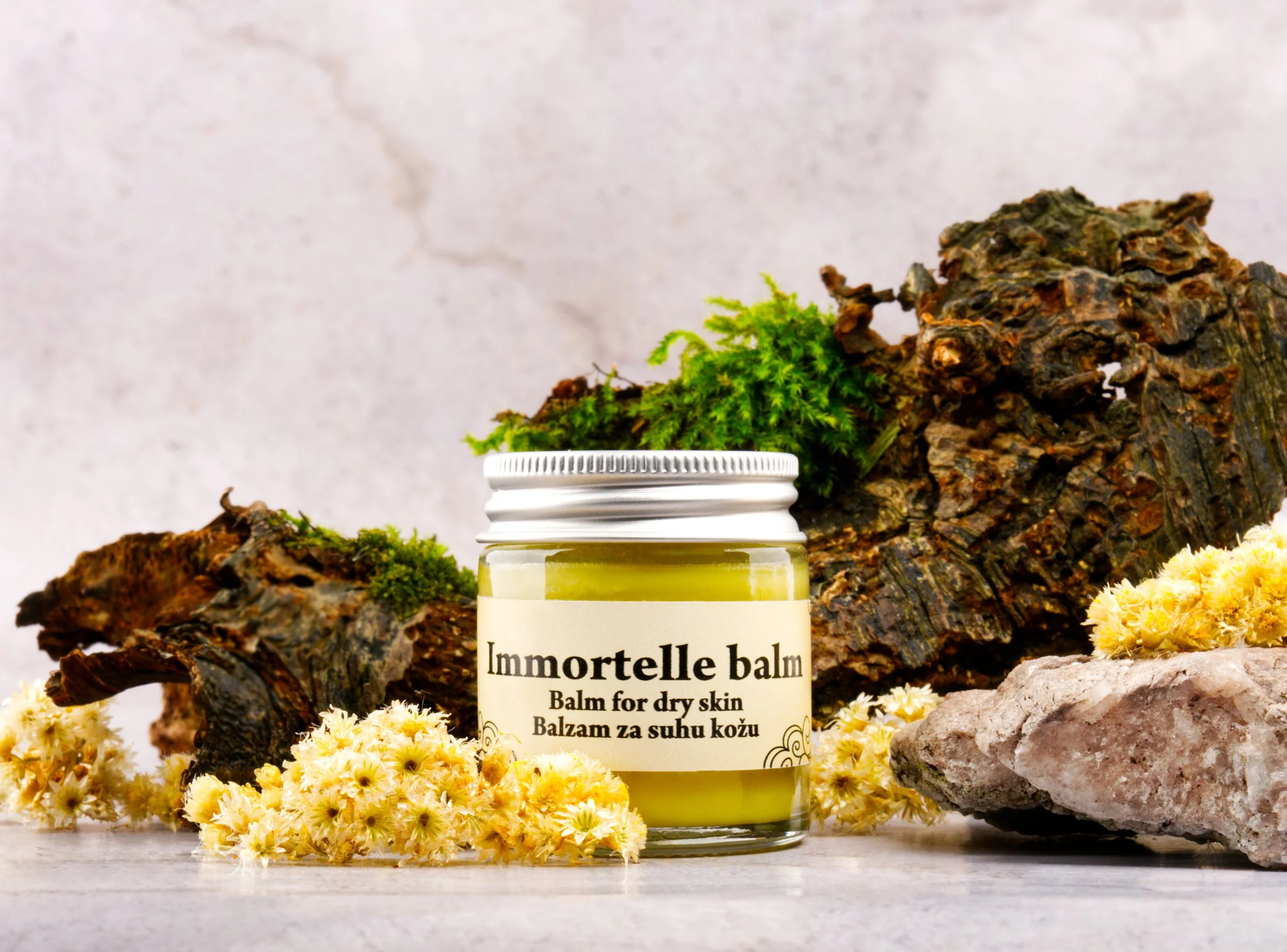 A jar of Immortelle balm surrounded by yellow flowers, green moss, and rocks on a light-colored surface with a marble background.