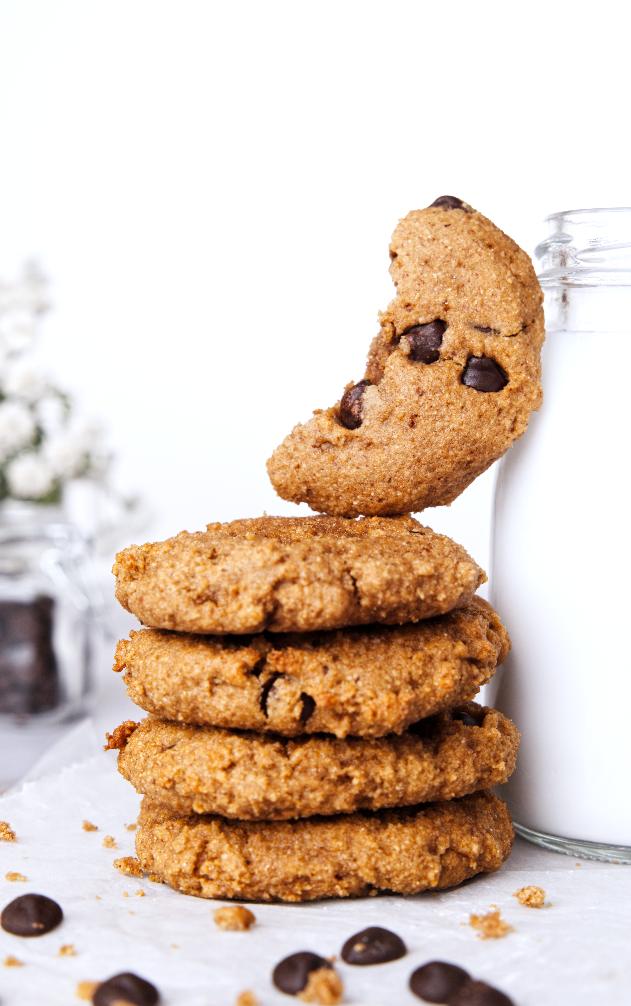 Stack of four chocolate chip cookies with a cookie on top and a glass jar of milk on the side, with scattered chocolate chips and crumbs around.