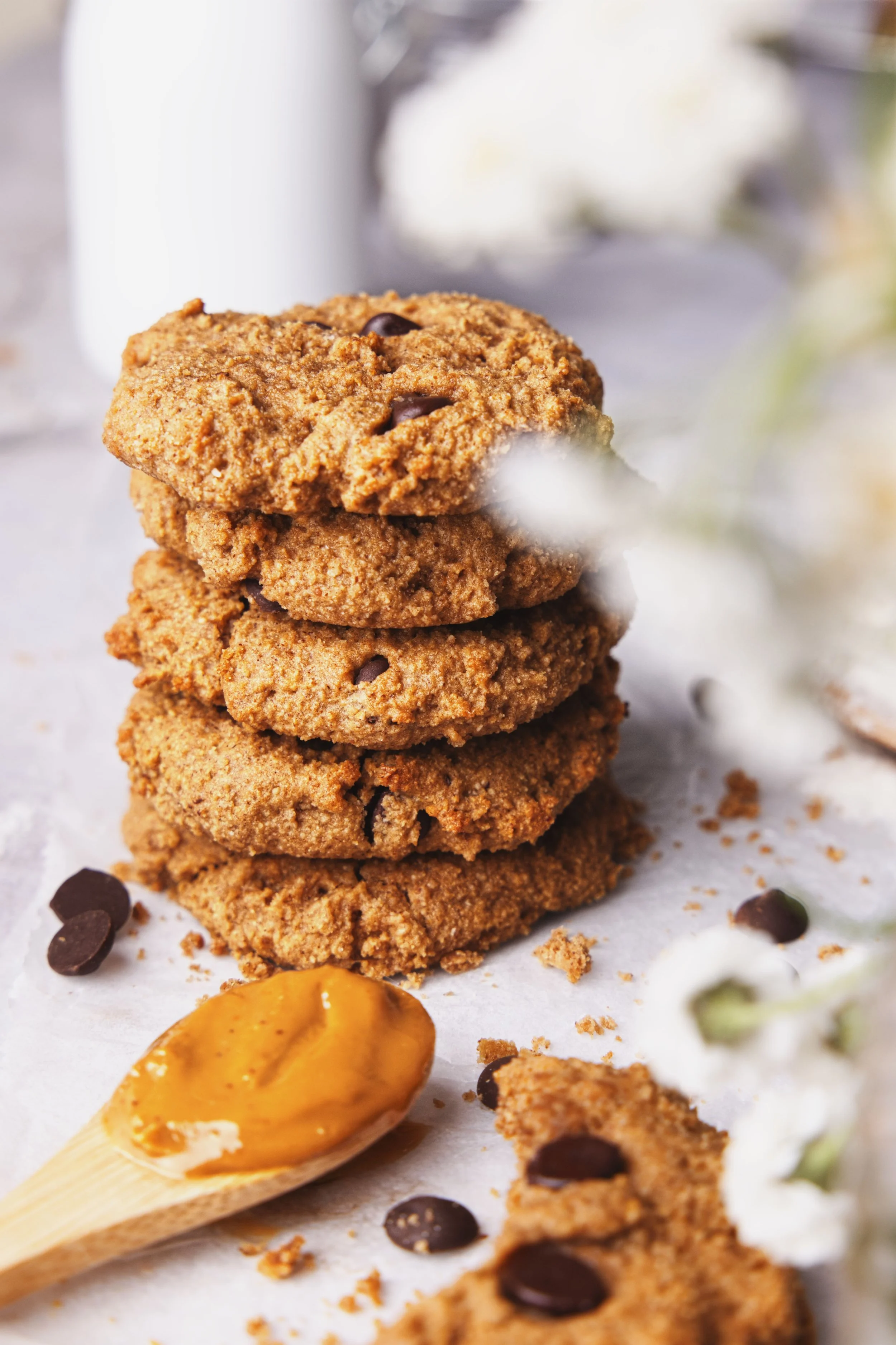 Stack of four chocolate chip cookies on white parchment paper, partially eaten, with scattered chocolate chips and a wooden spoon with peanut butter nearby.