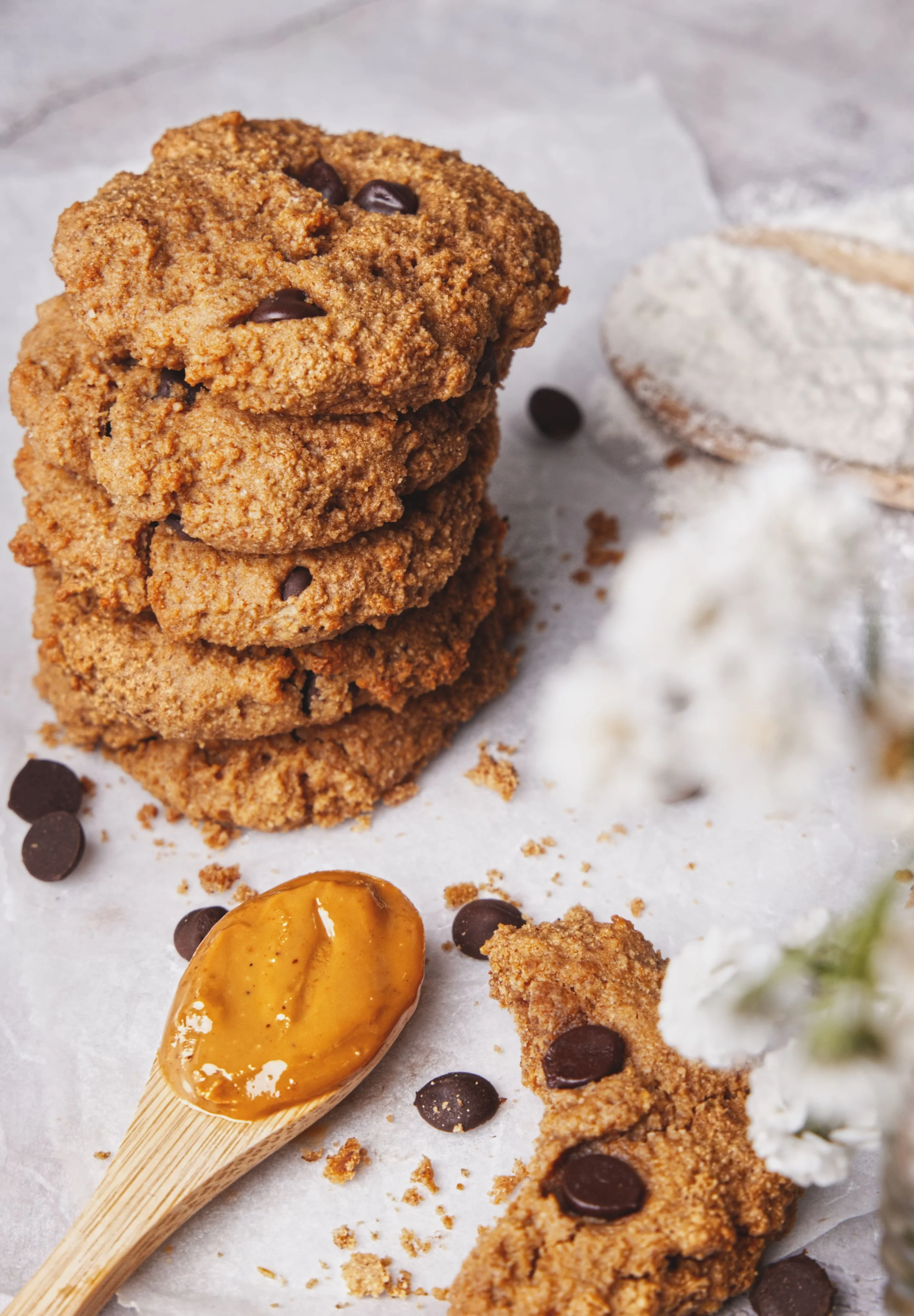 Close-up of three stacked oatmeal cookies with chocolate chips, a spoon with caramel sauce, and scattered chocolate chips on parchment paper.