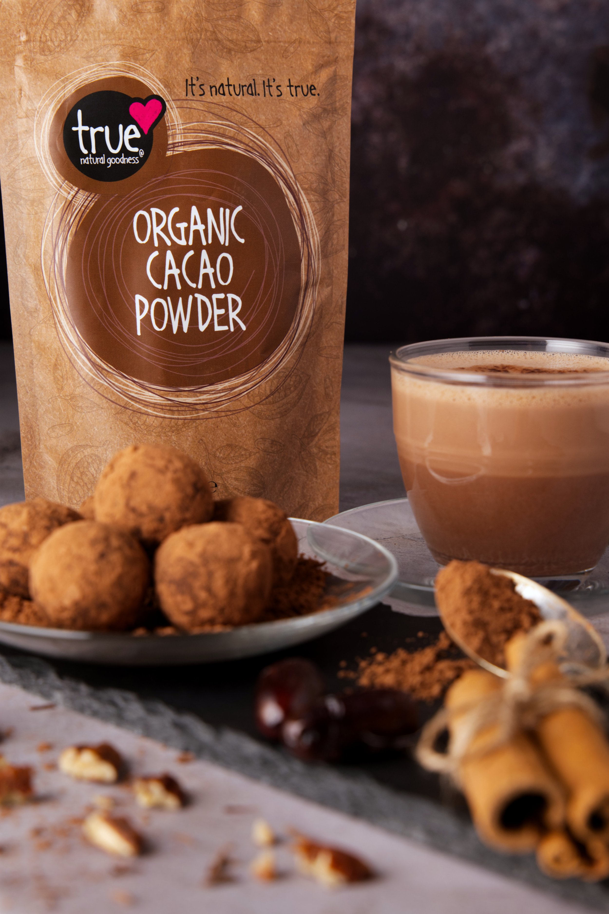 Close-up of a package of organic cacao powder, a glass of hot chocolate, a plate of chocolate truffles, and some broken cookies on a black slate surface.