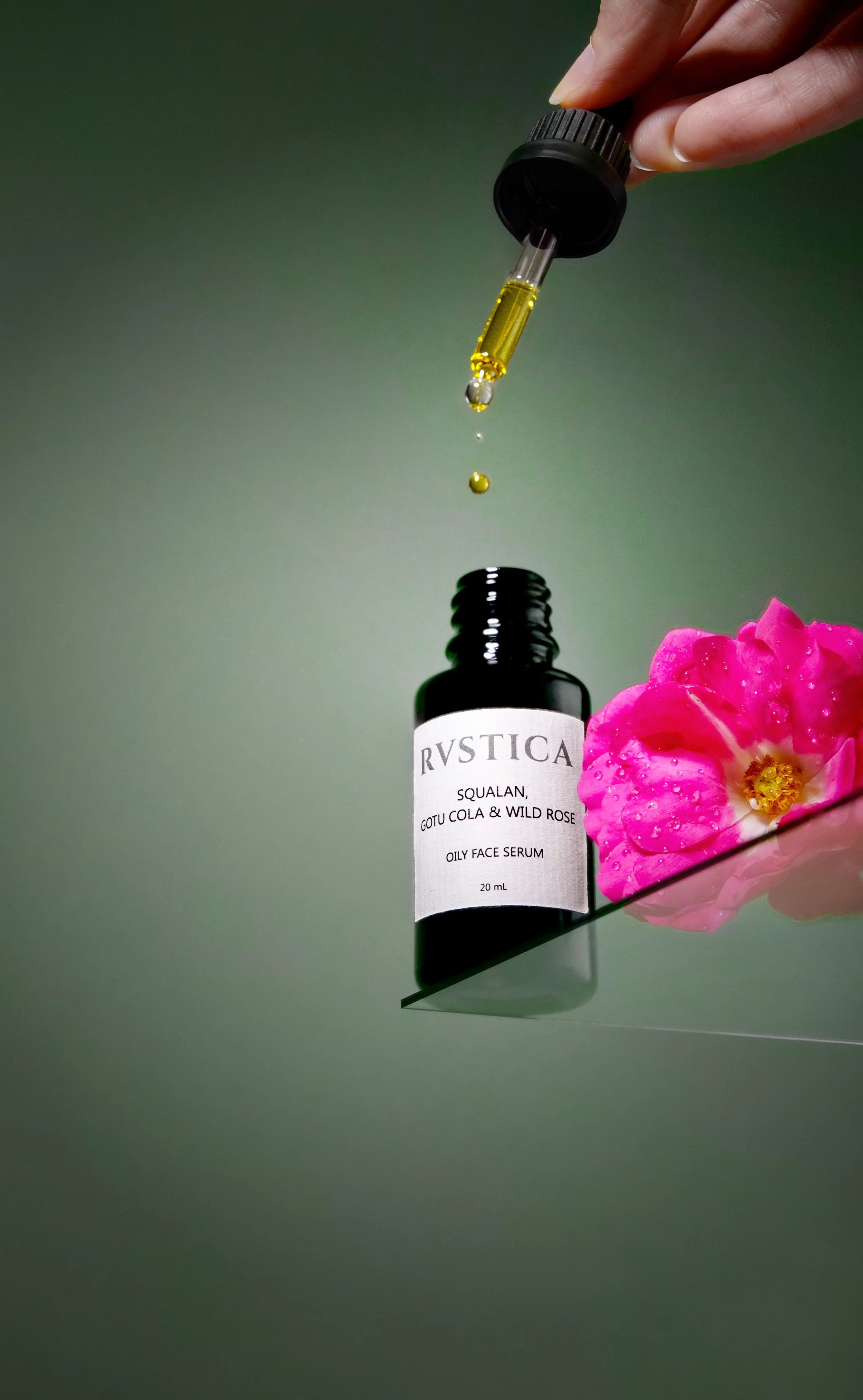 Close-up of a dropper with golden serum above a black bottle of skincare serum, with a pink flower next to the bottle on a reflective surface and green background.