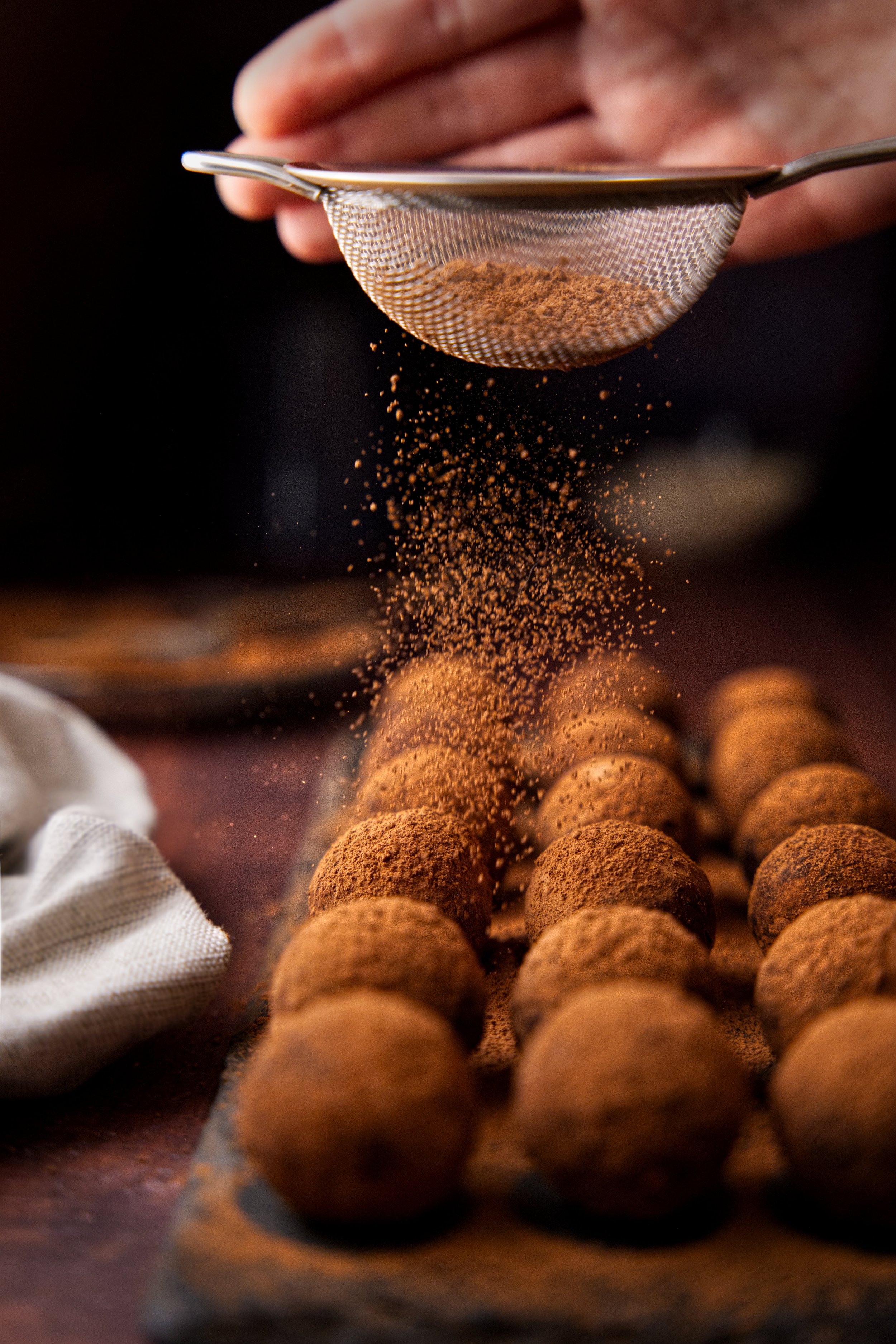 A close-up image of a person sifting cocoa powder over a line of round chocolate truffles on a dark surface.