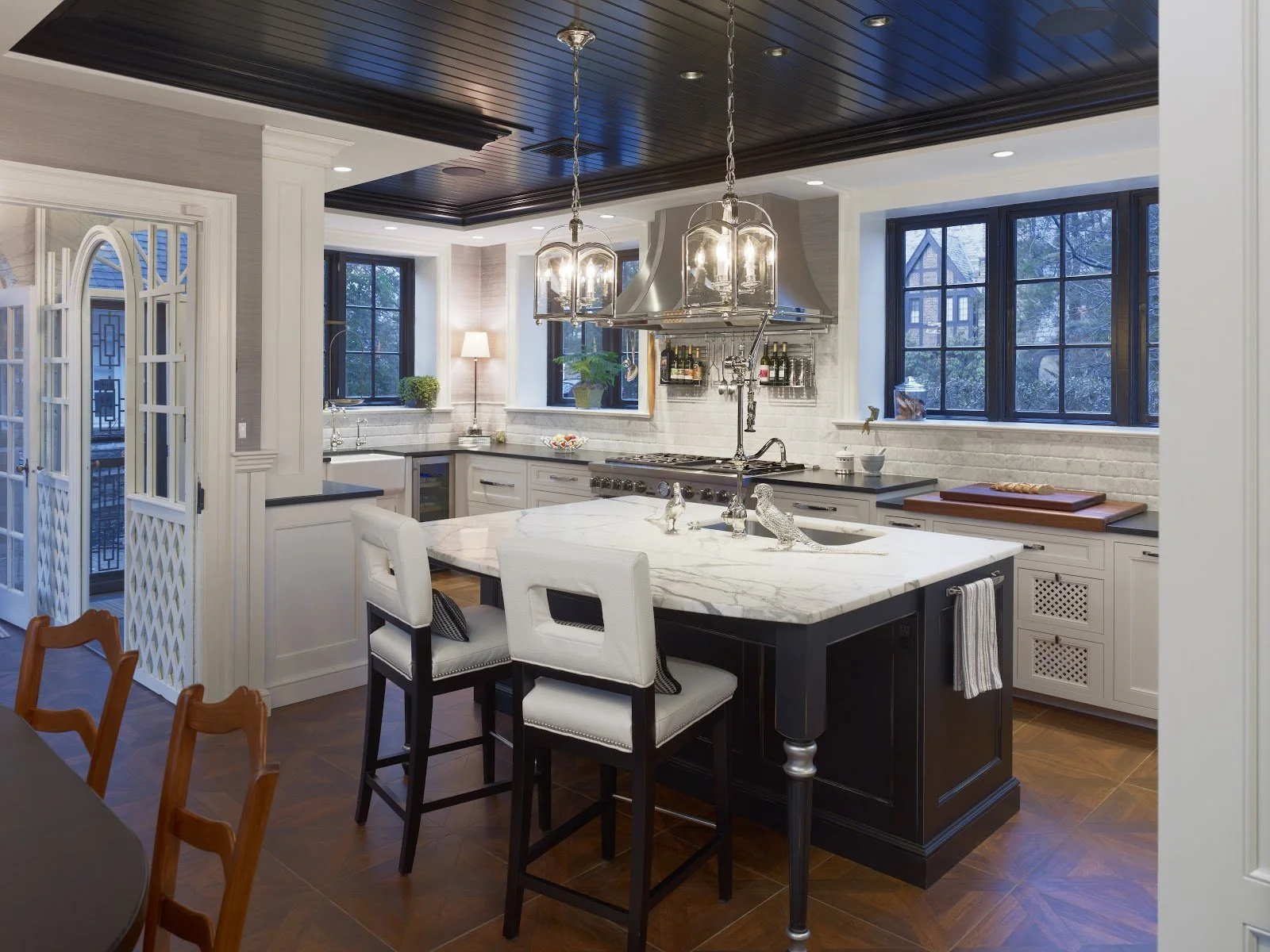Modern kitchen with white cabinets, black countertops, a marble island, dark wood ceiling, large windows, and stylish lighting fixtures.