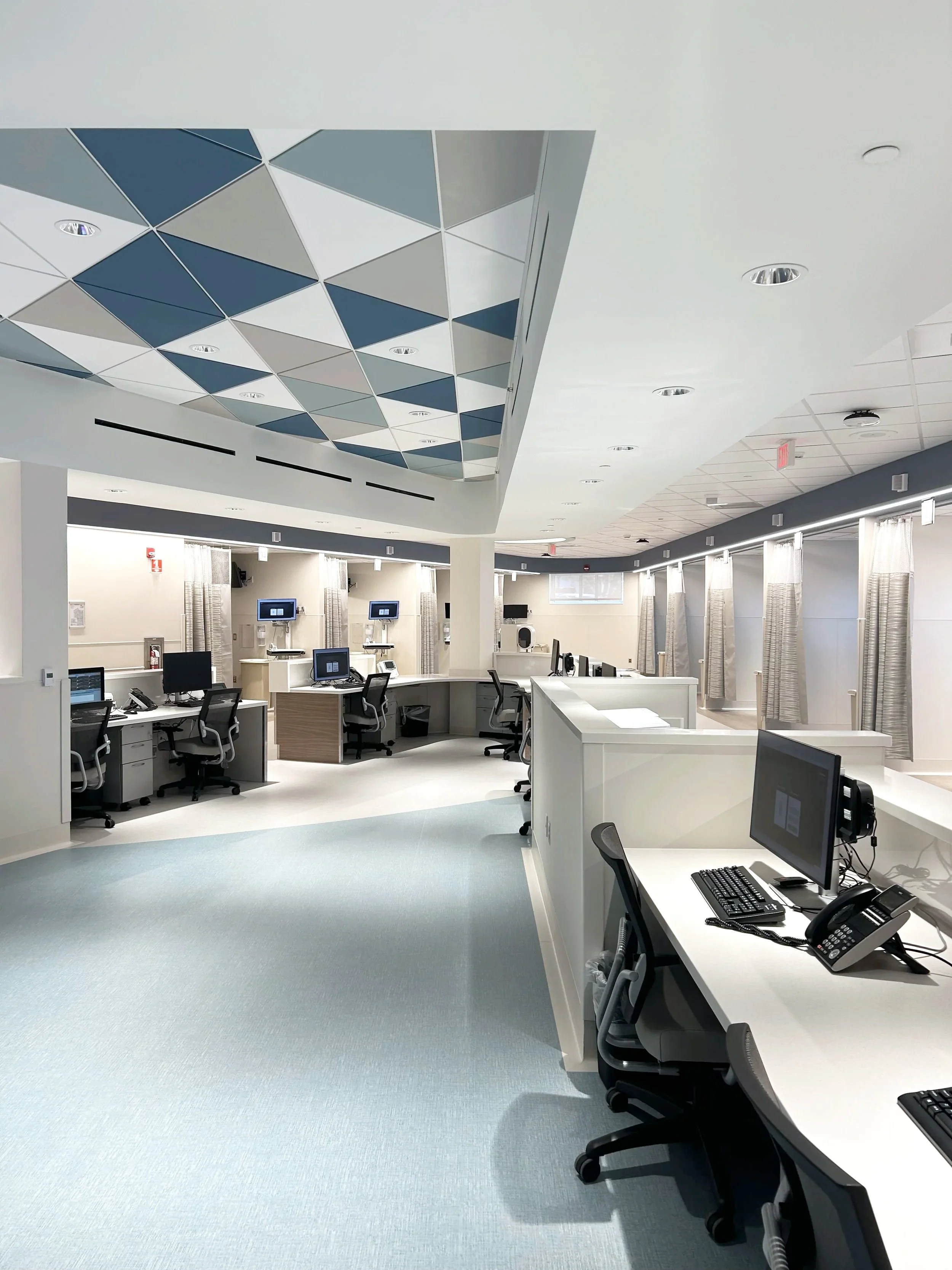Empty hospital nursing station with multiple workstations, computers, phones, and chairs, illuminated by overhead lights with a geometric ceiling design.