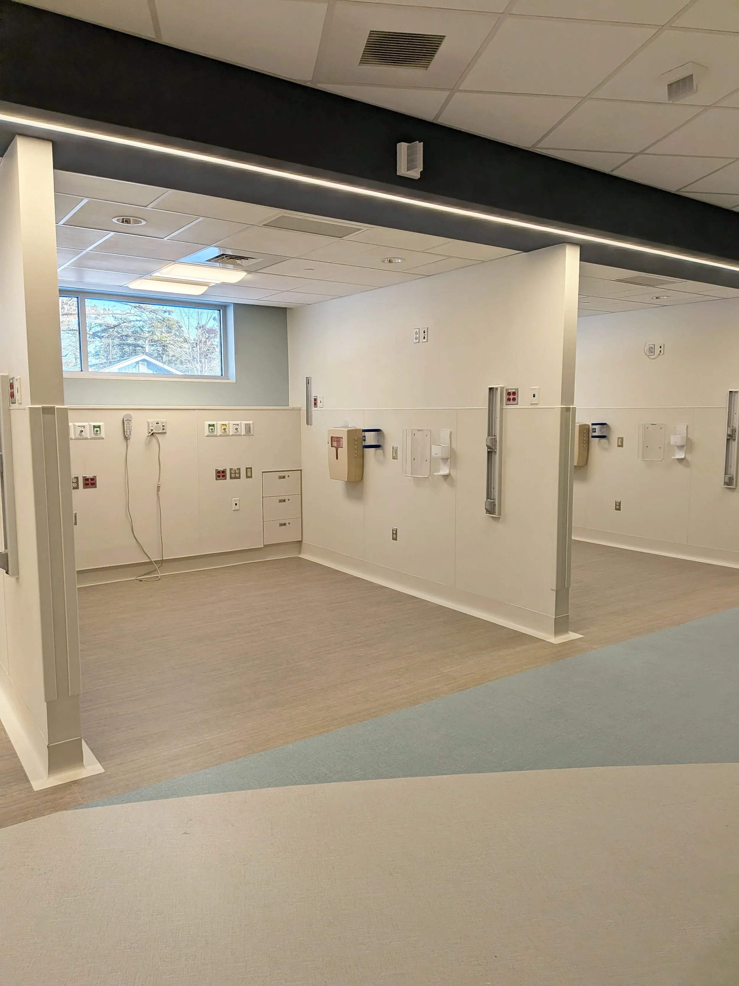 Empty hospital room with beige flooring, white walls, various medical outlets and equipment, and a window showing outdoor trees and a house.