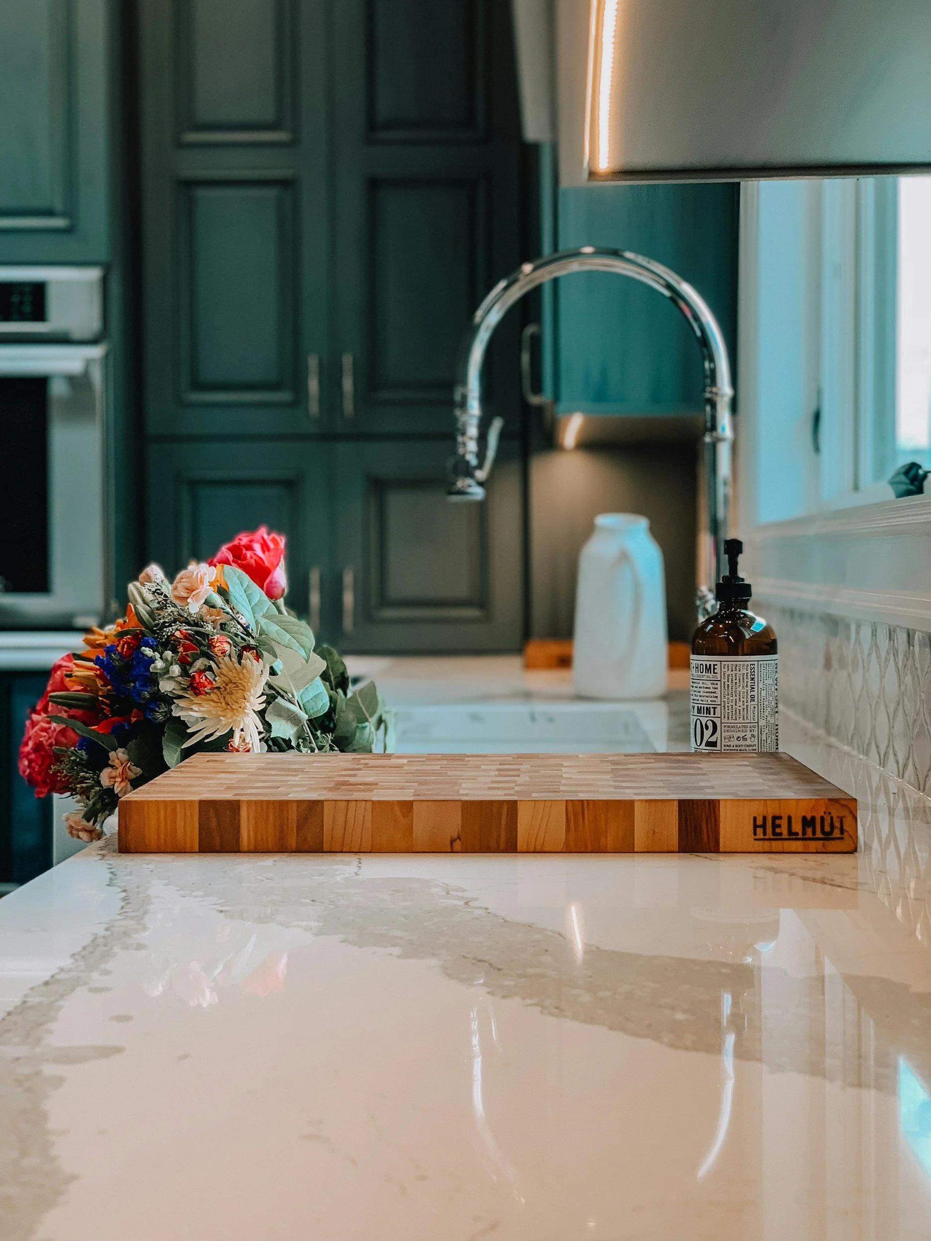 A kitchen countertop with a wooden HELMÜT cutting board, a white and brown soap dispenser, and a vase with colorful flowers, with dark green cabinets in the background.