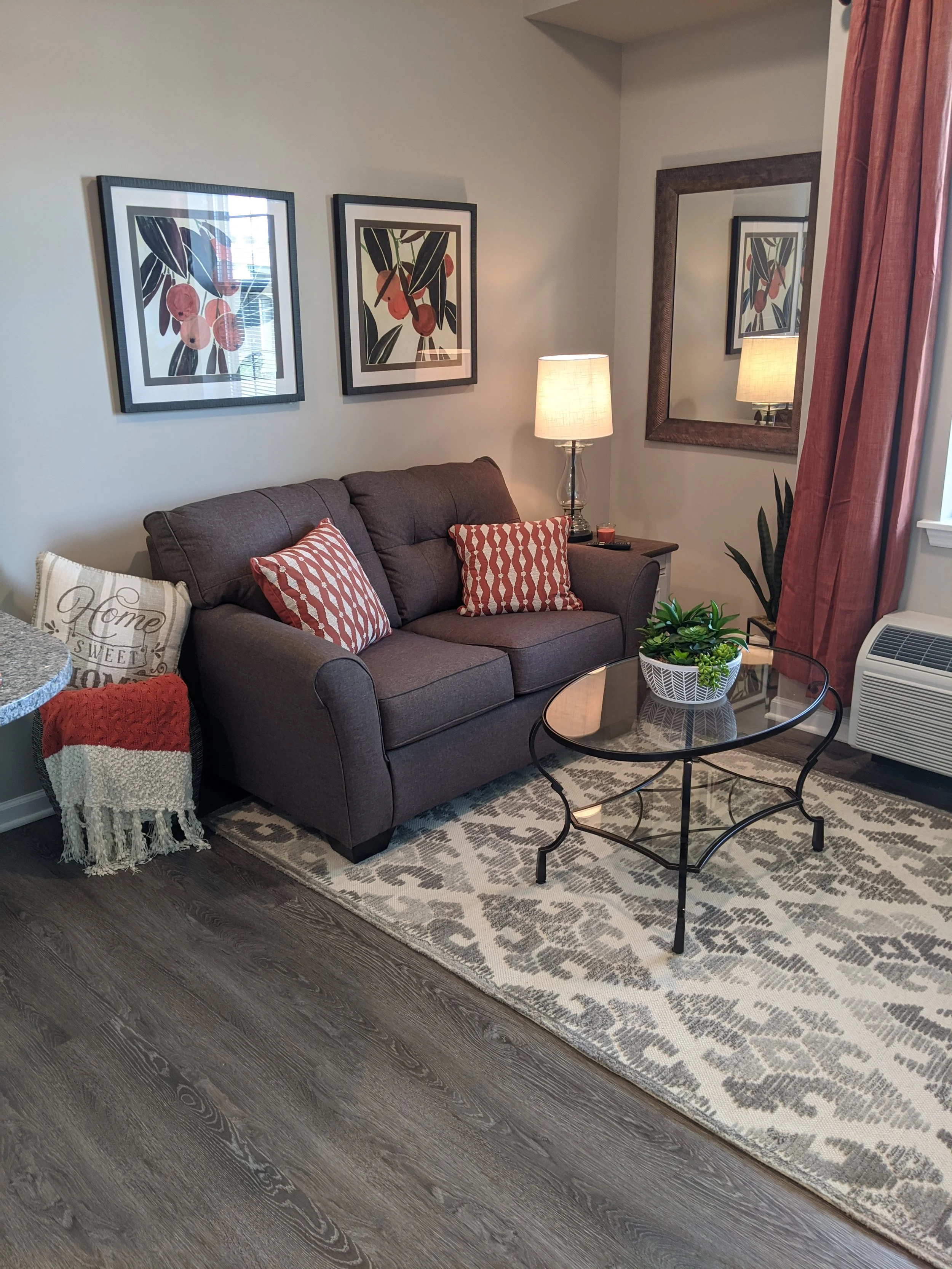 A living room with a gray sofa with red and beige patterned pillows, a beige and white throw blanket, a small wooden side table with a glass lamp, framed artwork of orange fruits and green leaves on the wall, a large mirror, a window with red curtain