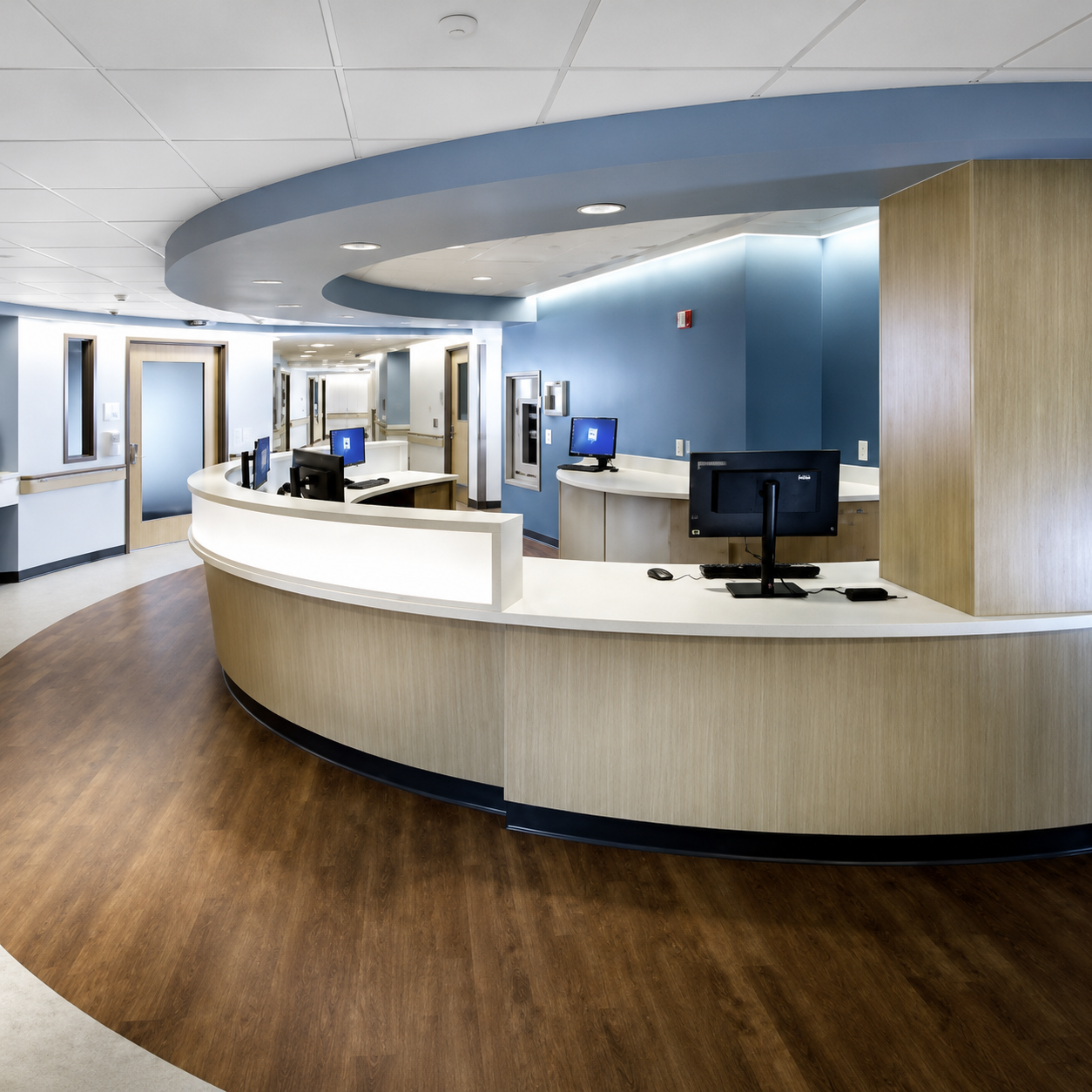 Hospital reception area with curved wooden desk, computers, blue walls, and ceiling lighting.