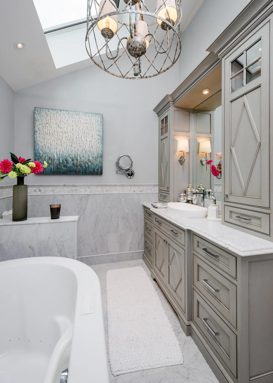A bright bathroom with a skylight, a chandelier, a gray vanity with a white marble countertop, a mirror, a white bathtub, a black vase with pink and white flowers, and abstract artwork on the wall.