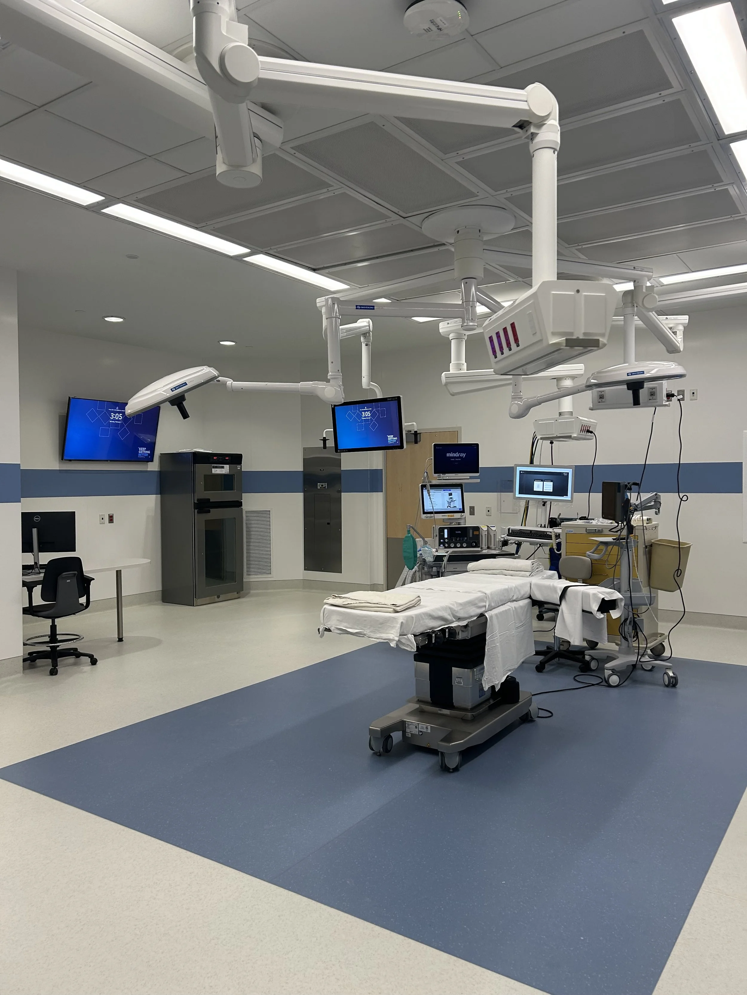 Hospital operating room with surgical table, medical equipment, multiple monitors, and overhead lights.