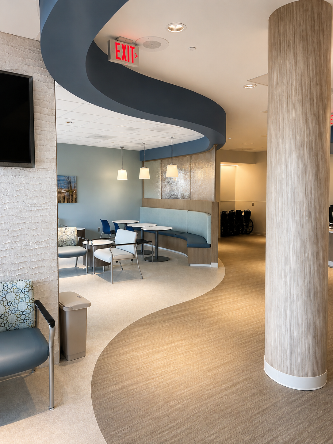 Empty indoor waiting area with chairs, tables, pendant lights, and a curved ceiling feature in a medical or office building.