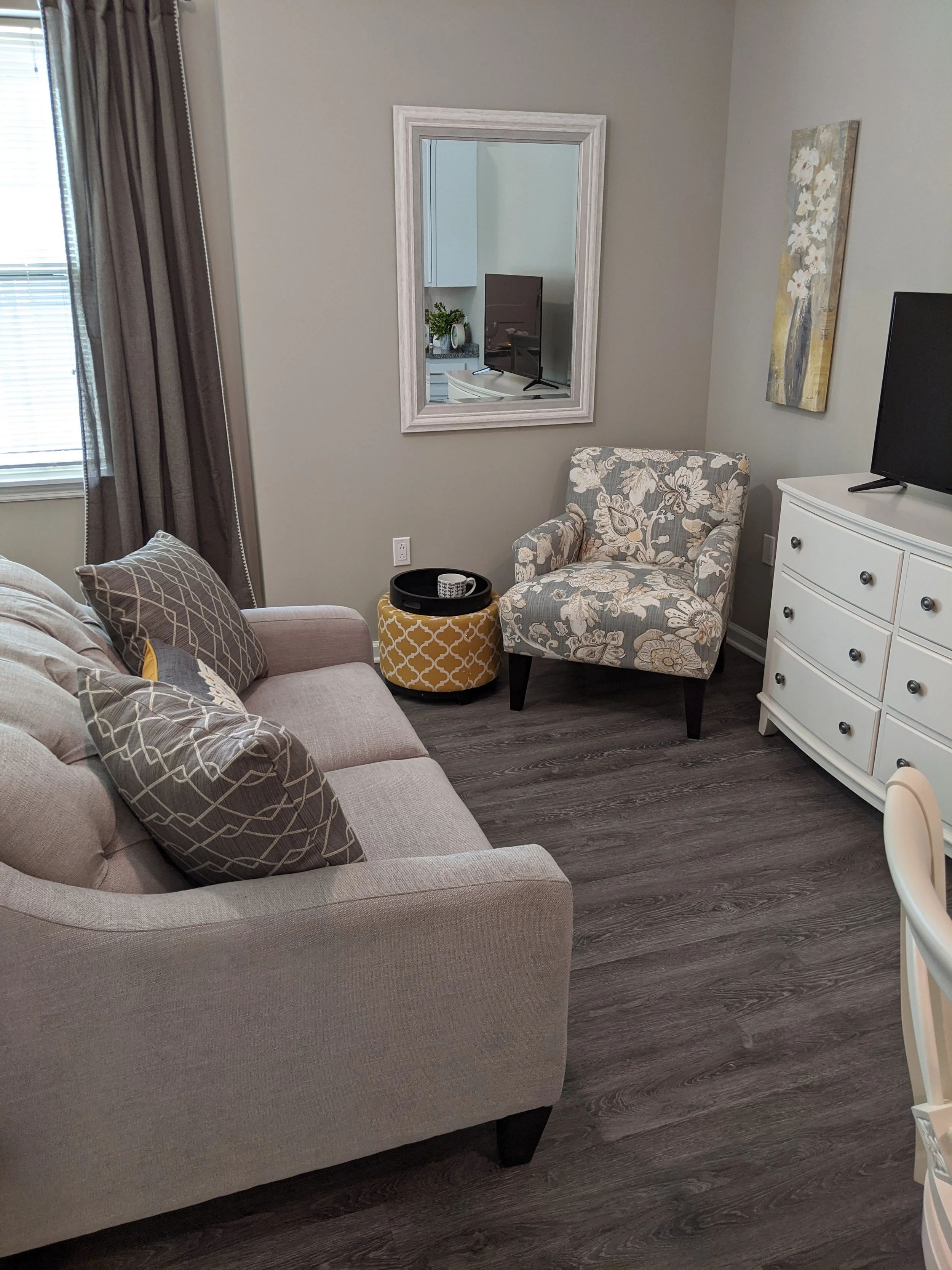 Living room corner with a beige sofa, patterned pillows, a floral patterned armchair, a white dresser with a TV, a black round tray with a mug, artwork on the wall, a mirror reflecting kitchen cabinets, and window with brown curtains.