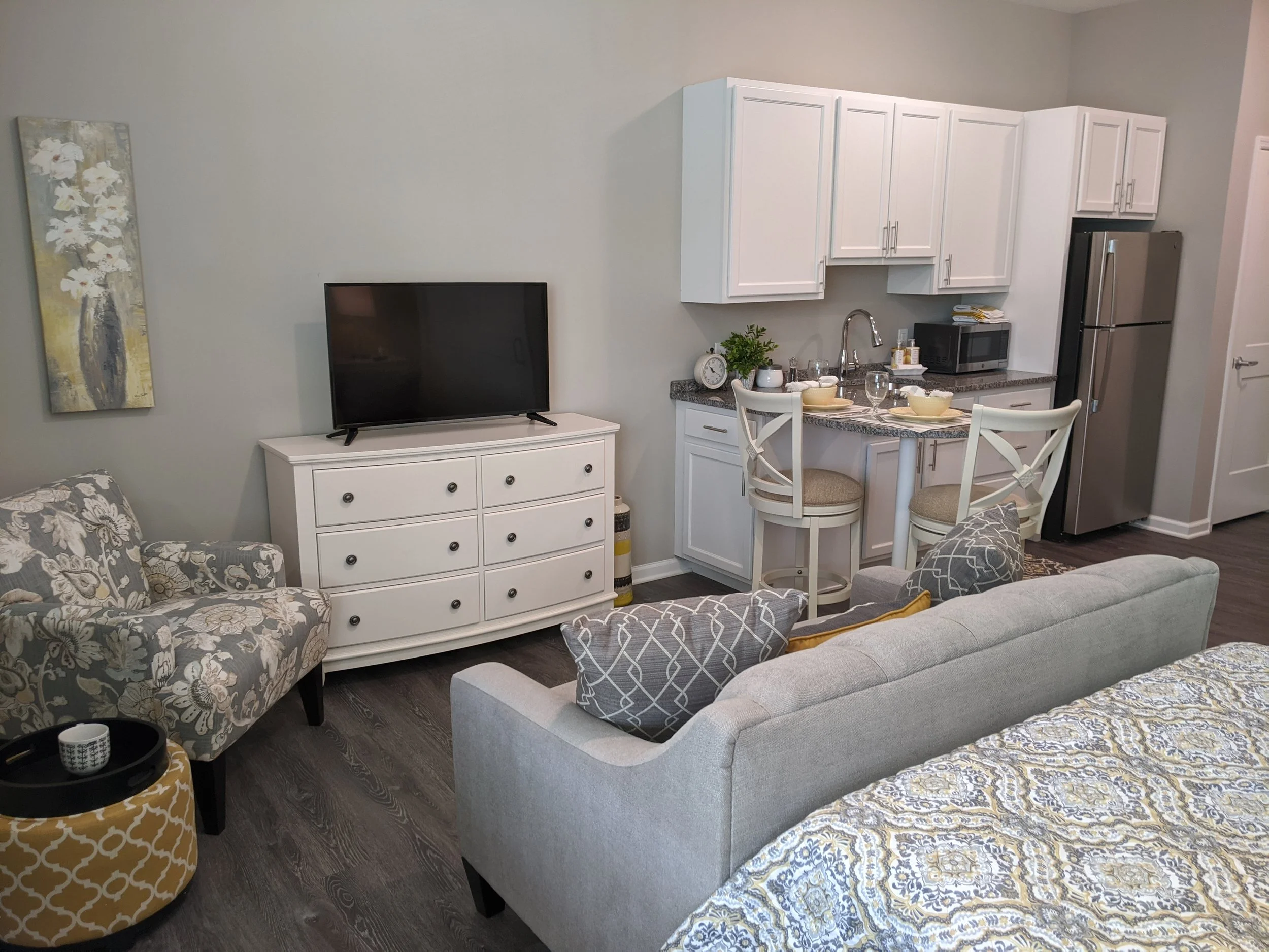 Living room with a gray sofa, floral patterned armchair, white media console with a TV, kitchen area with white cabinets, stainless steel refrigerator, dining table with chairs, and wall art.