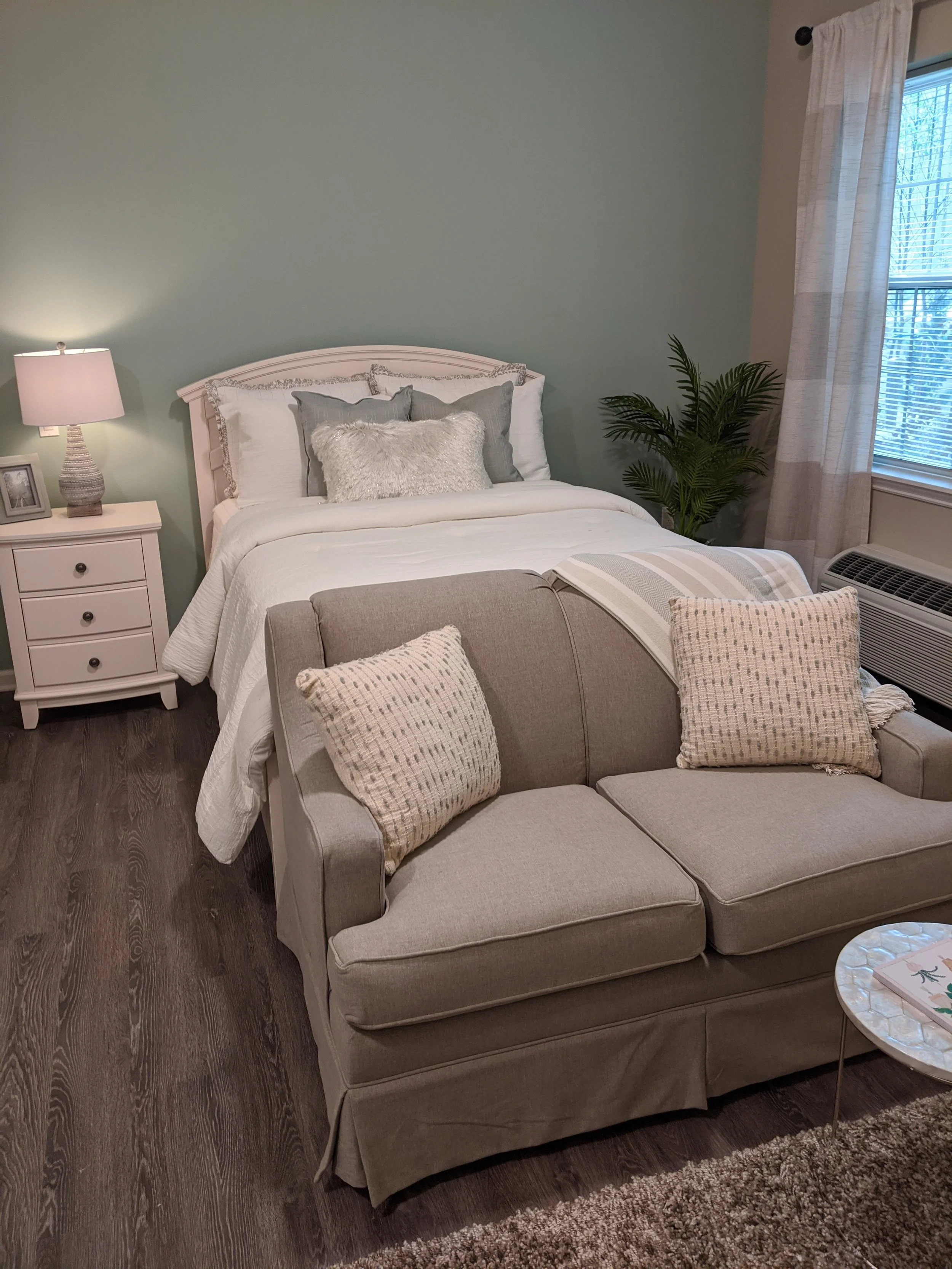 A bedroom with a white bed, beige and gray pillows, a white nightstand with a lamp, a picture frame, a green plant, and a window with cream curtains, dark wood flooring, and a beige upholstered armchair with plaid pillows in front of the bed.