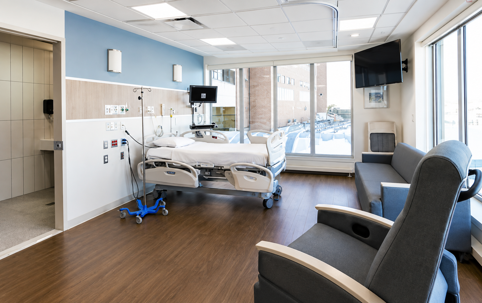 Hospital room with a hospital bed, medical equipment, a TV mounted on the wall, a window with a view outside, a gray sofa, and chairs.