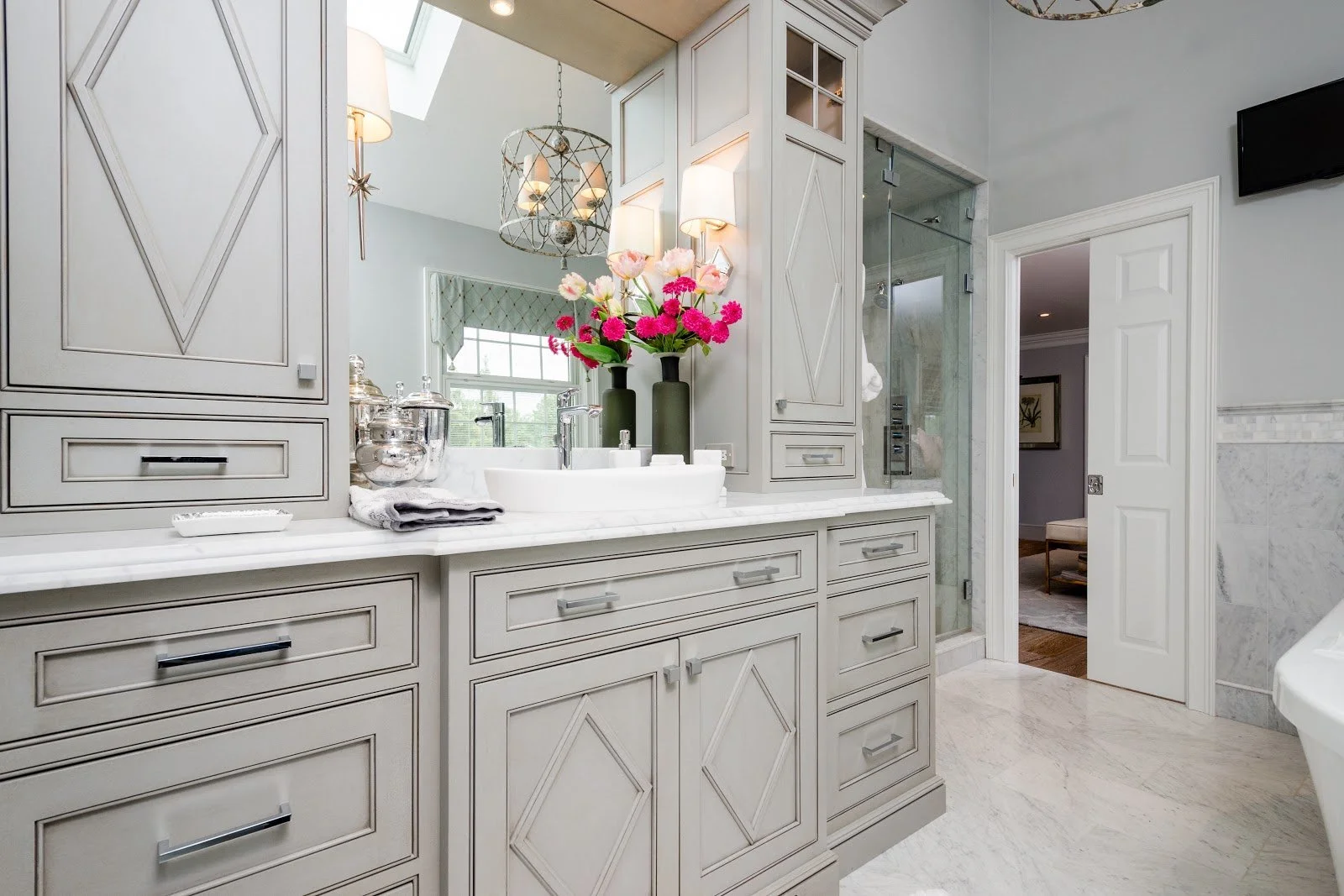 A luxurious bathroom with a gray vanity, a marble countertop, and two vases with pink and white flowers. There is a large mirror, a shower enclosure, and a door leading to another room. The bathroom has white walls and ceiling with modern lighting fi