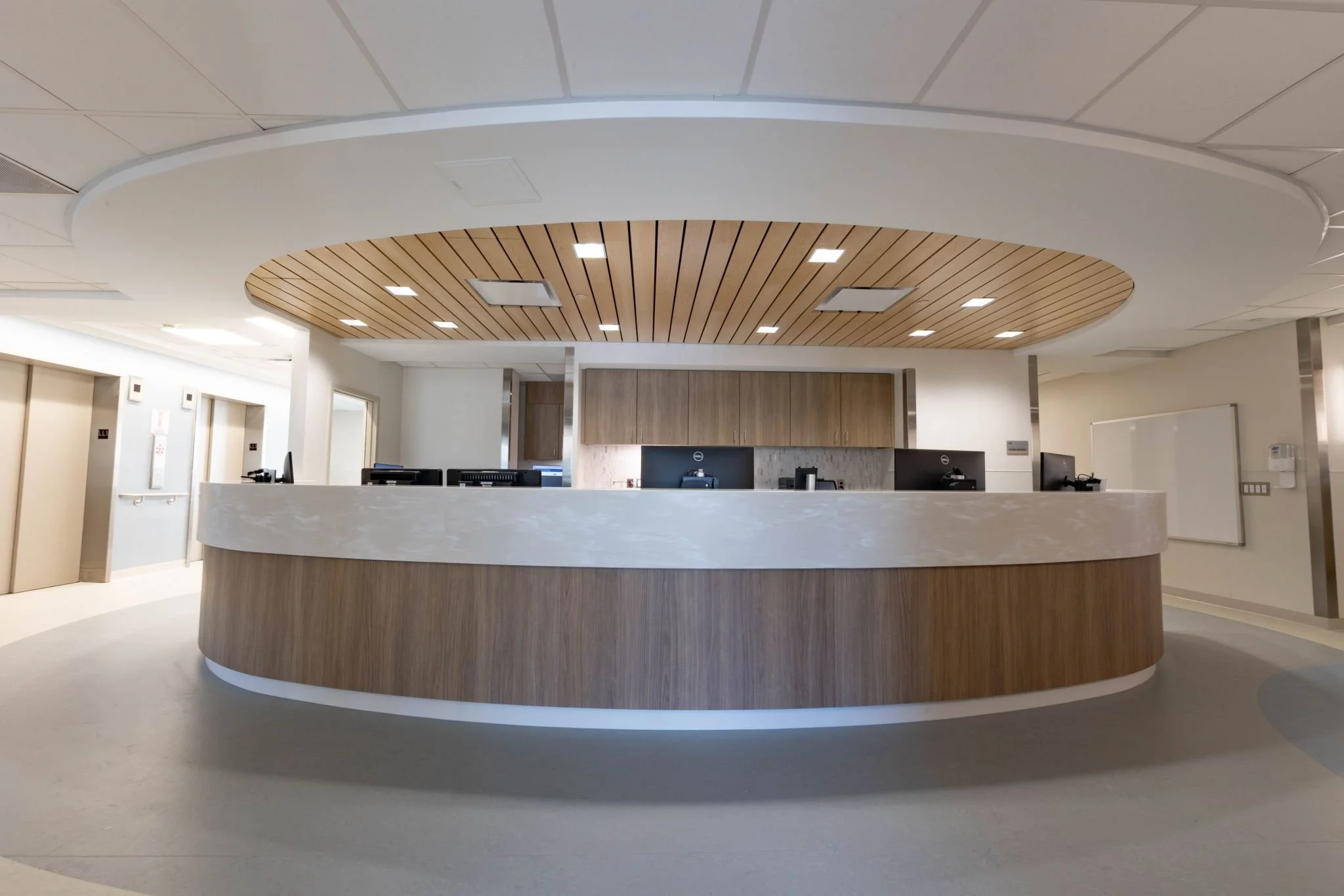 A modern hospital or office reception area with a circular wooden reception desk, ceiling with wood paneling, and multiple computer stations behind the desk.