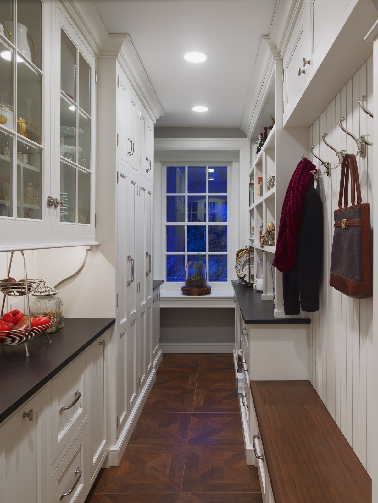 A narrow laundry or mudroom with white cabinetry, a large window at the end, and a wooden bench with coat hooks on the right wall.