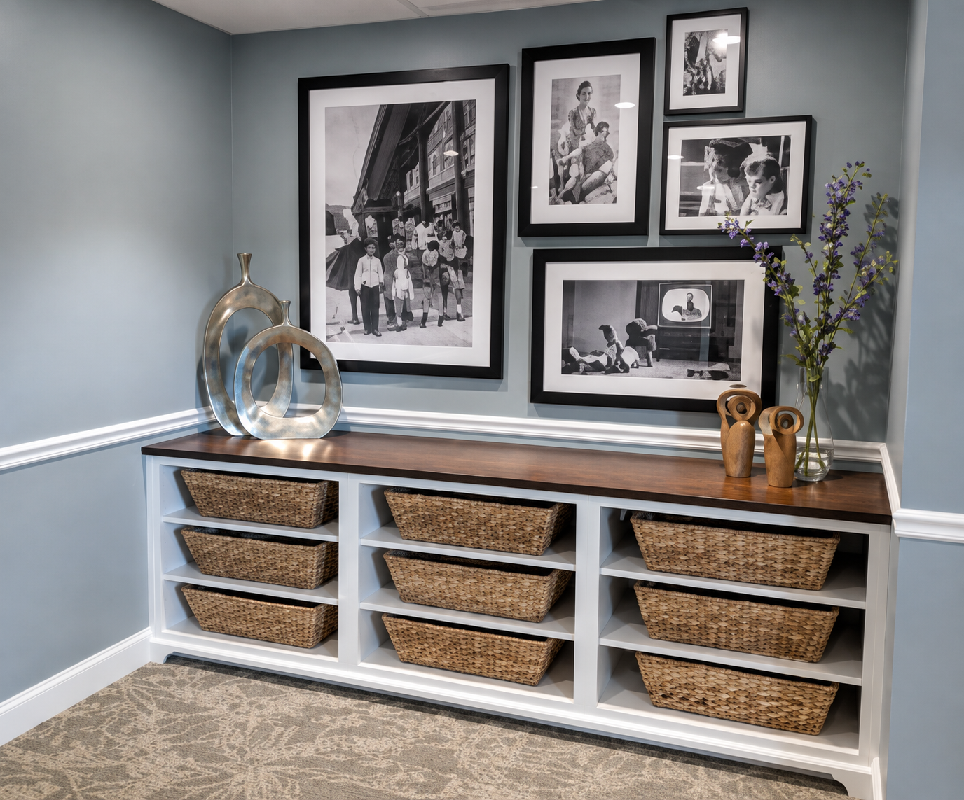 A wall decorated with six black and white framed photographs, a wooden sideboard with woven baskets, and decorative vases and flowers on top.