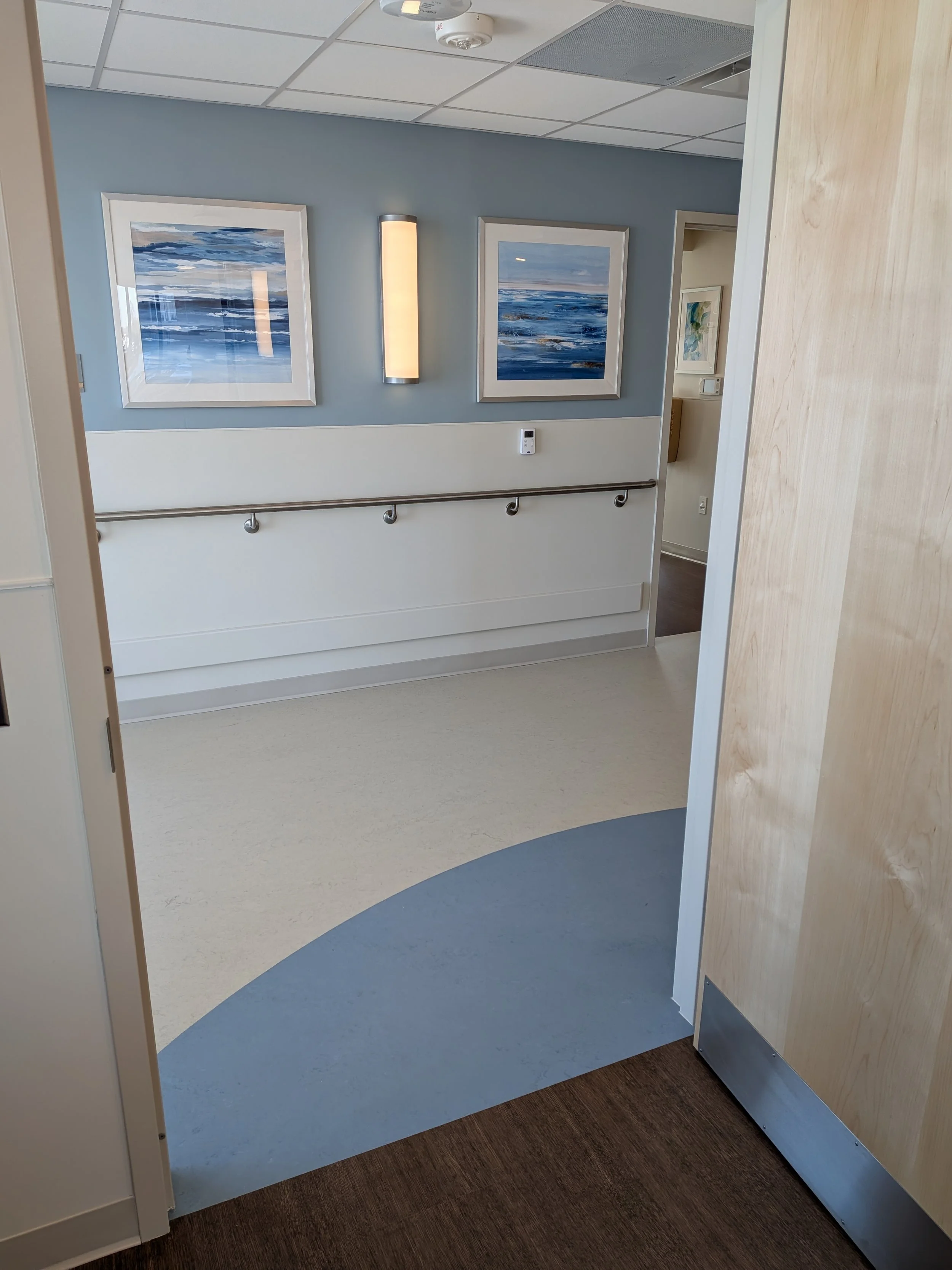 Hospital corridor with blue and beige flooring, light blue walls, framed ocean-themed artwork, and a wall-mounted light fixture.