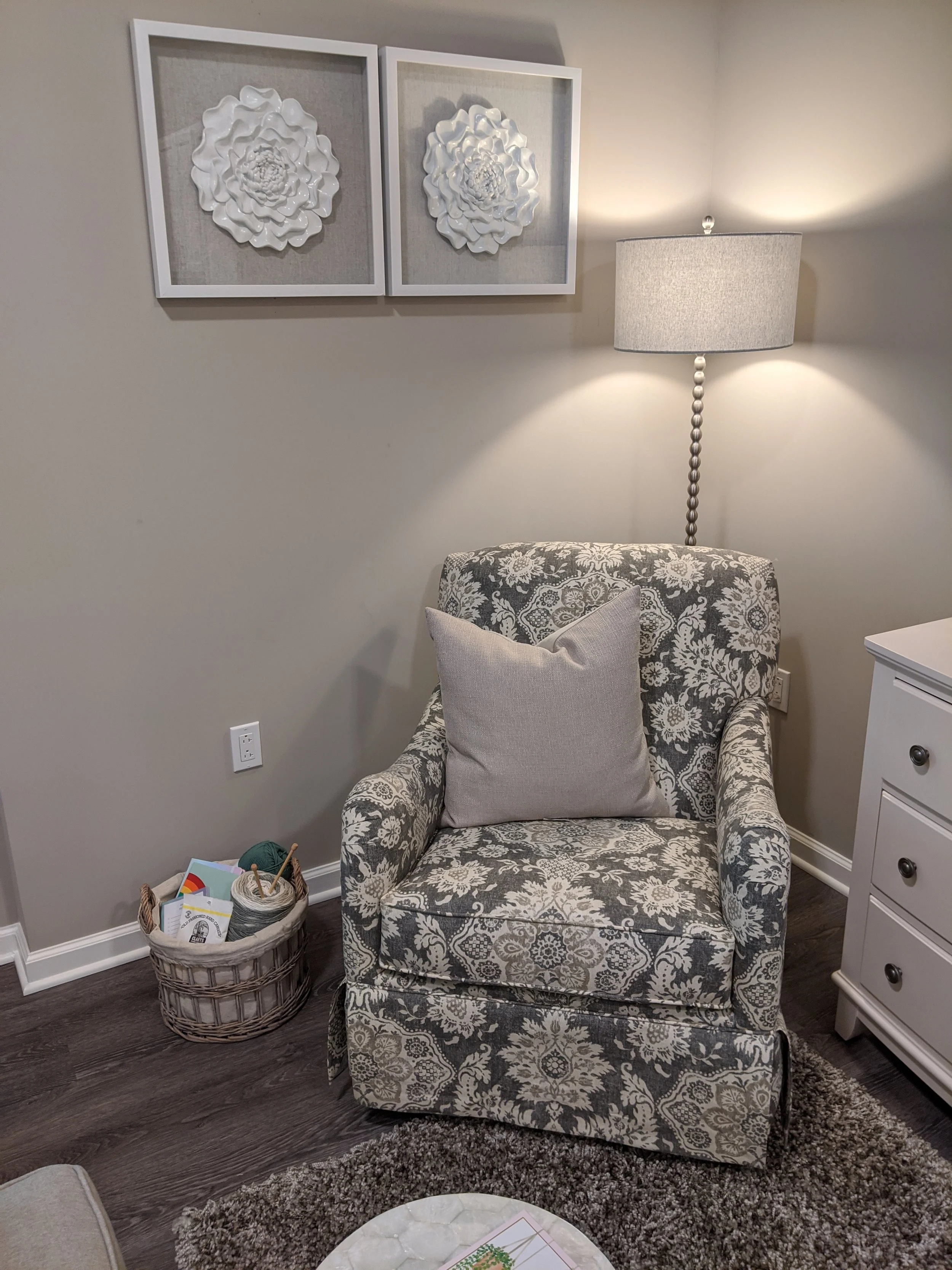 A cozy corner with a patterned armchair, a beige pillow, a white dresser, and a basket filled with yarn and knitting accessories. A tall floor lamp with a beige lampshade provides lighting. Two framed 3D floral wall art pieces hang above the chair, a