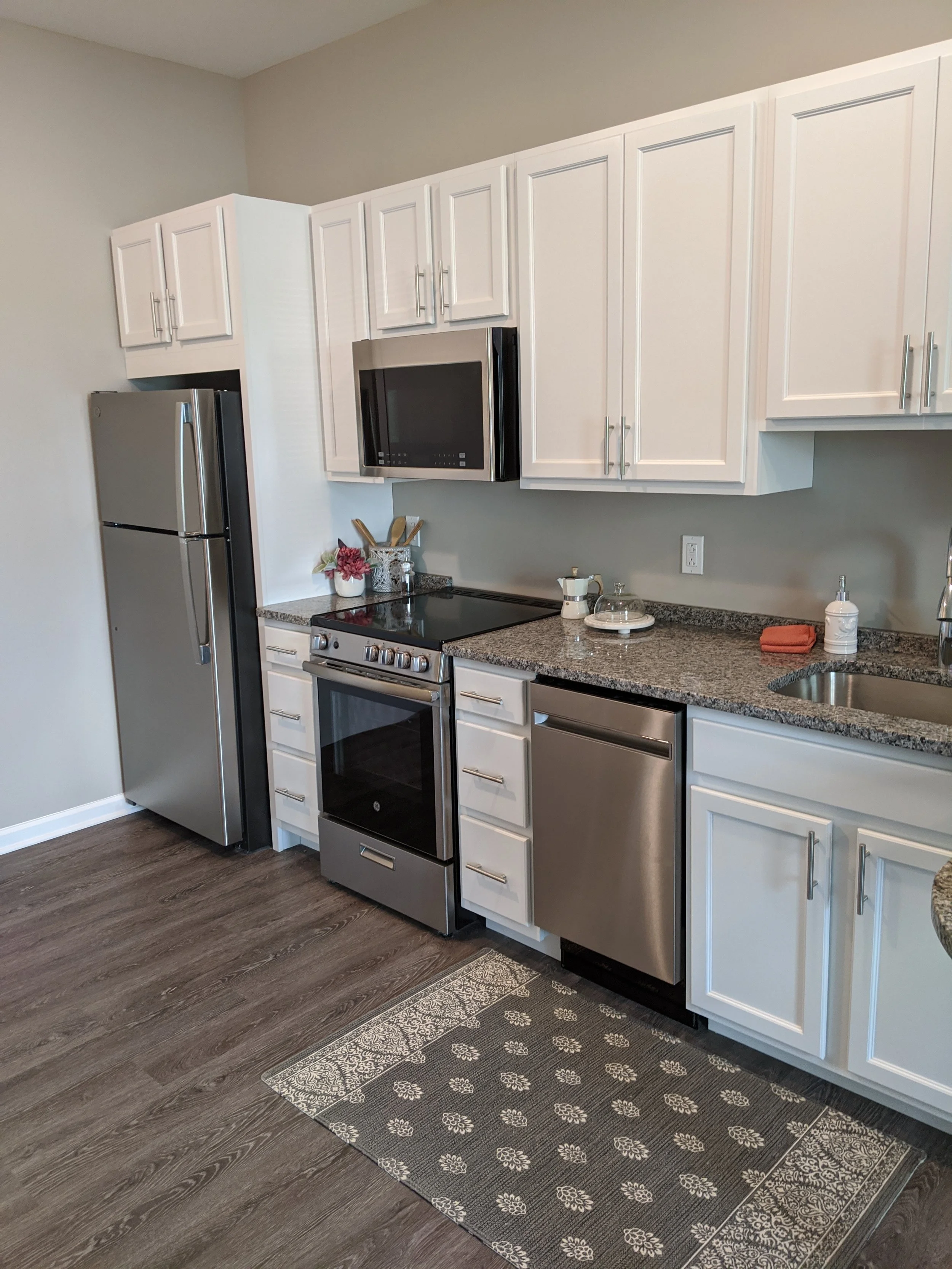 Kitchen with white cabinets, stainless steel refrigerator, stove, microwave, granite countertops, and a patterned rug on dark wood flooring.