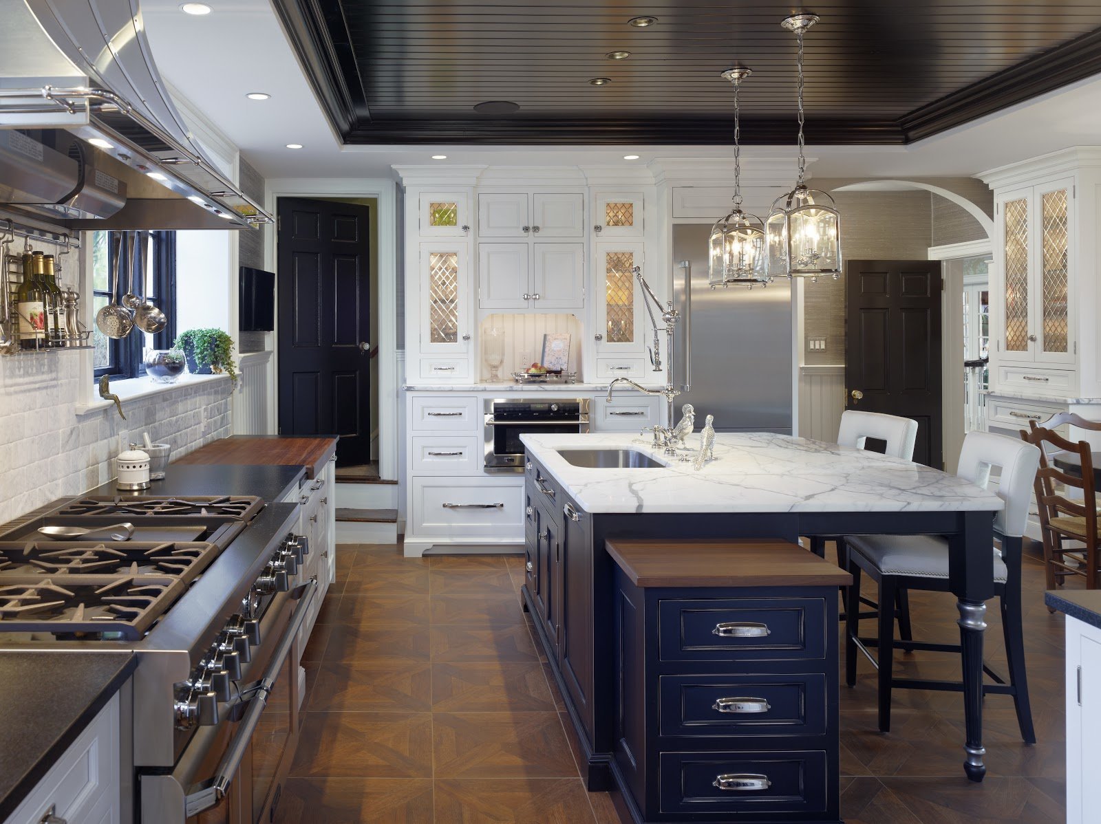 A spacious, modern kitchen with white cabinetry, a dark wood ceiling, a marble island, stainless steel appliances, and black accents, illuminated by chandelier lights.