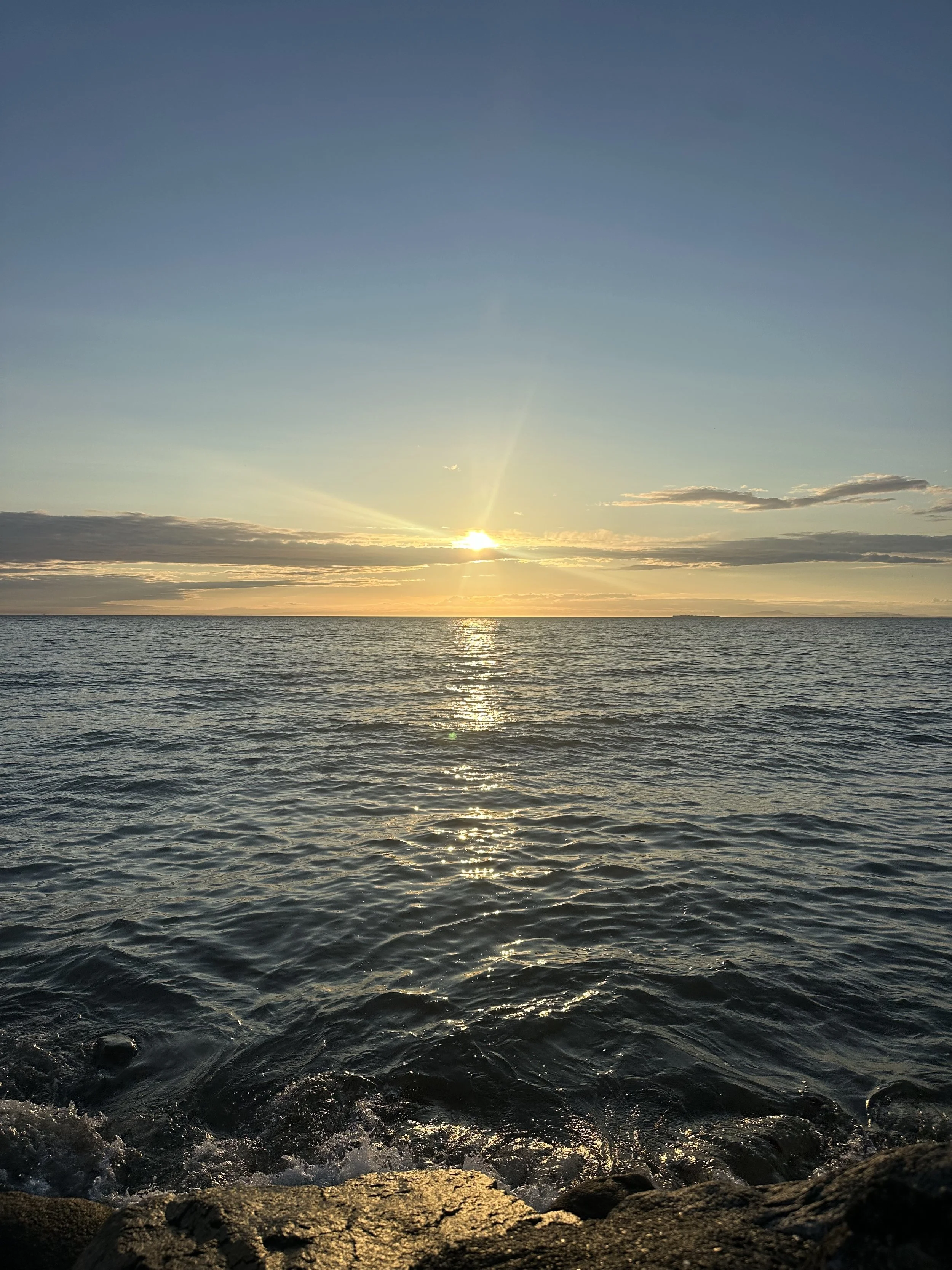 Sunset over a calm ocean with reflections on the water, rocky shoreline in the foreground, and a partly cloudy sky.