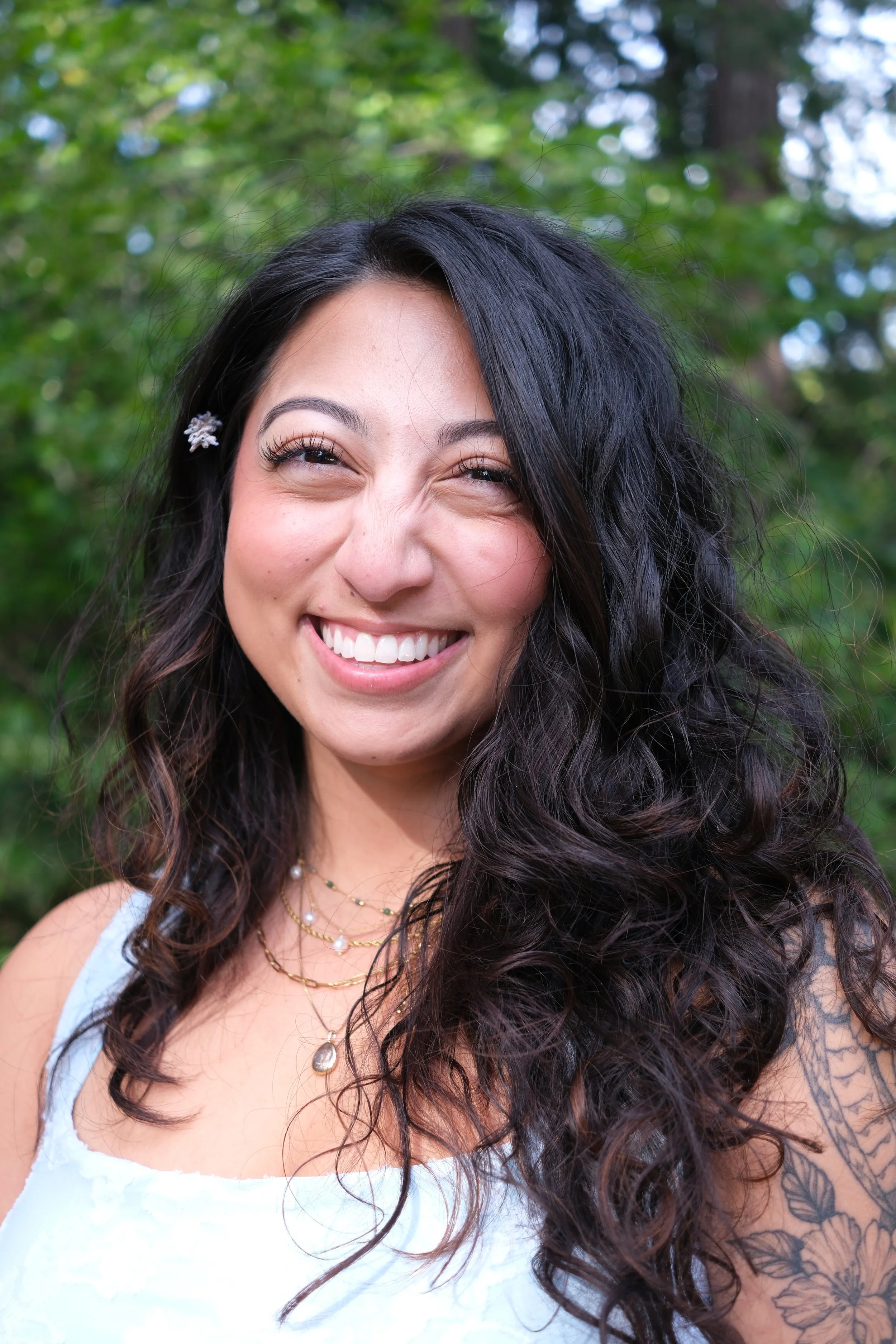 Close-up of a smiling woman with long, curly dark hair, outdoors with green trees in the background.