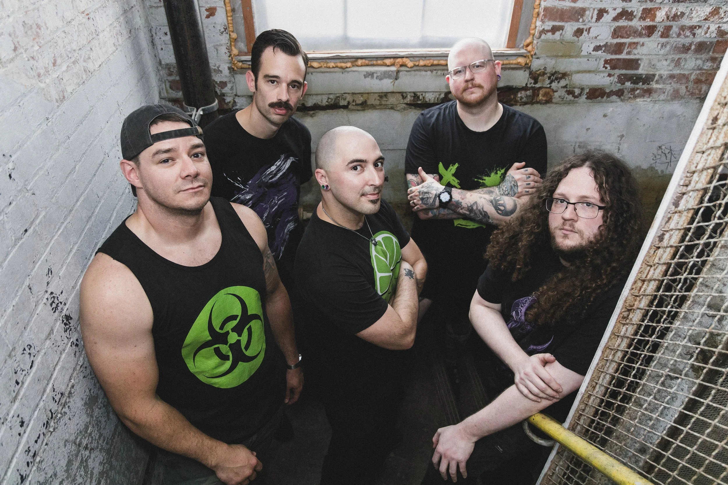A group of five men standing in a small, distressed indoor space with exposed brick and paint, some with tattoos and piercings, all with serious expressions.