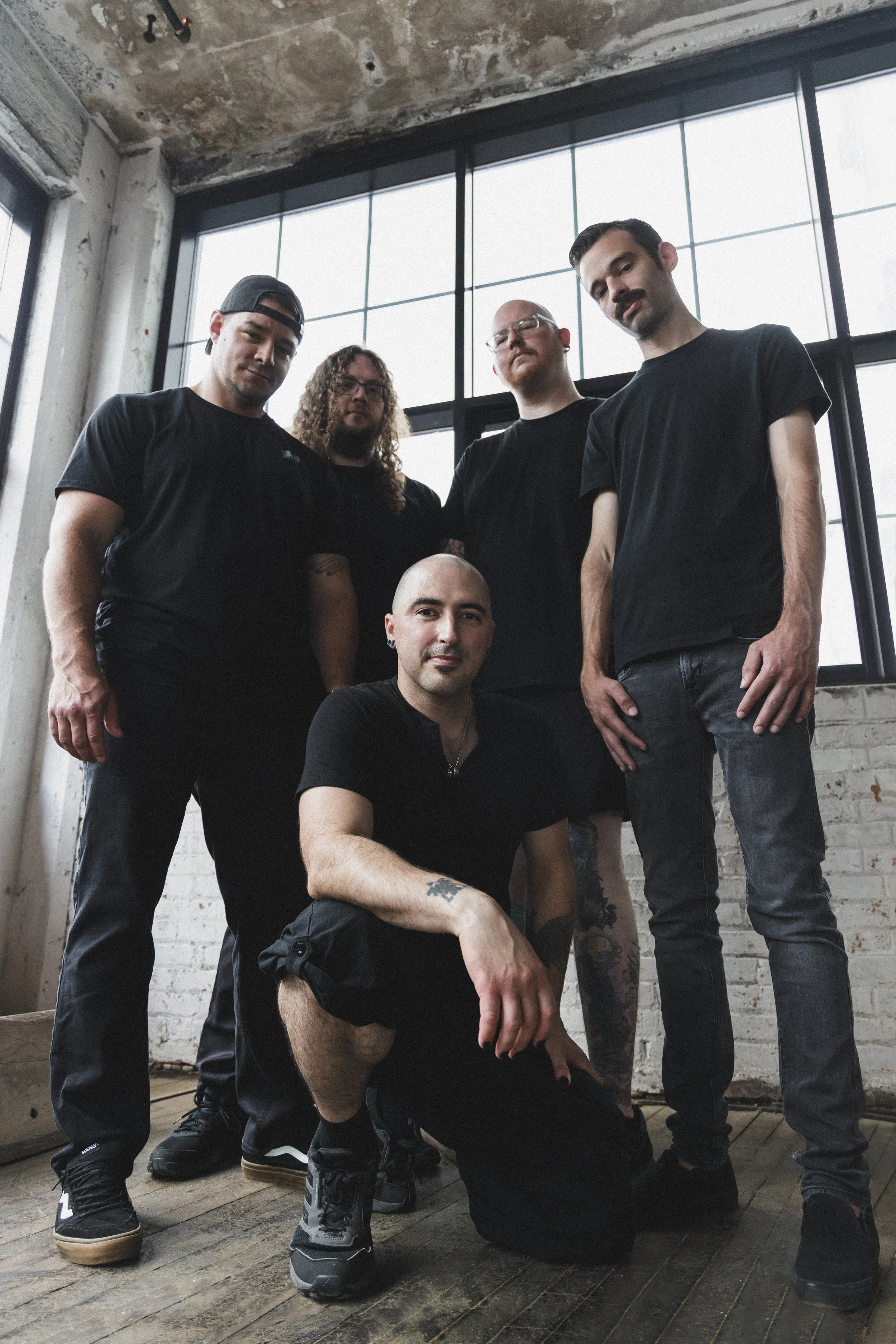 Five men in black clothing posing in an industrial-style room with large windows and a wooden floor, one kneeling and four standing.