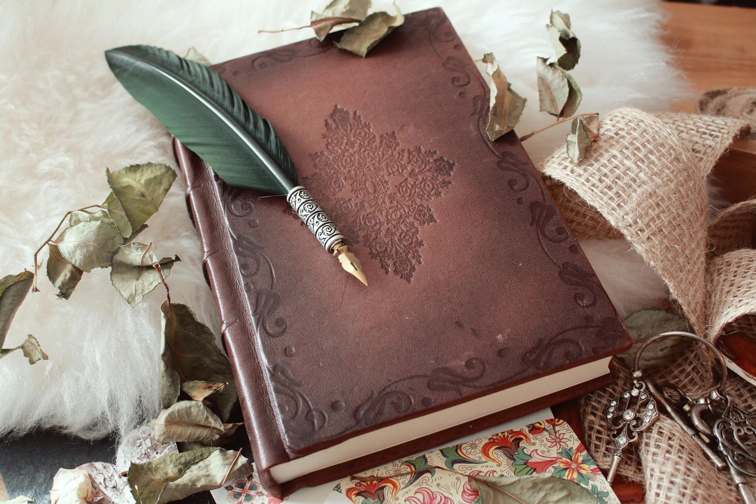 Un cuaderno de cuero marrón con un diseño en relieve en la tapa, acompañando una pluma estilográfica con detalles en blanco y negro y varios objetos decorativos, rodeados de ramas secas y otros artículos decorativos.