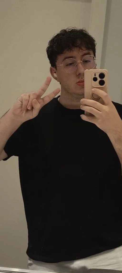 Selfie Dan Zaga, chico joven con pelo rizado y oscuro, con gafas redondas con borde color oro rosa, movil con funda oro rosa y cascos en las orejas de color oro rosa. Camiseta negra y pose con dos dedos.