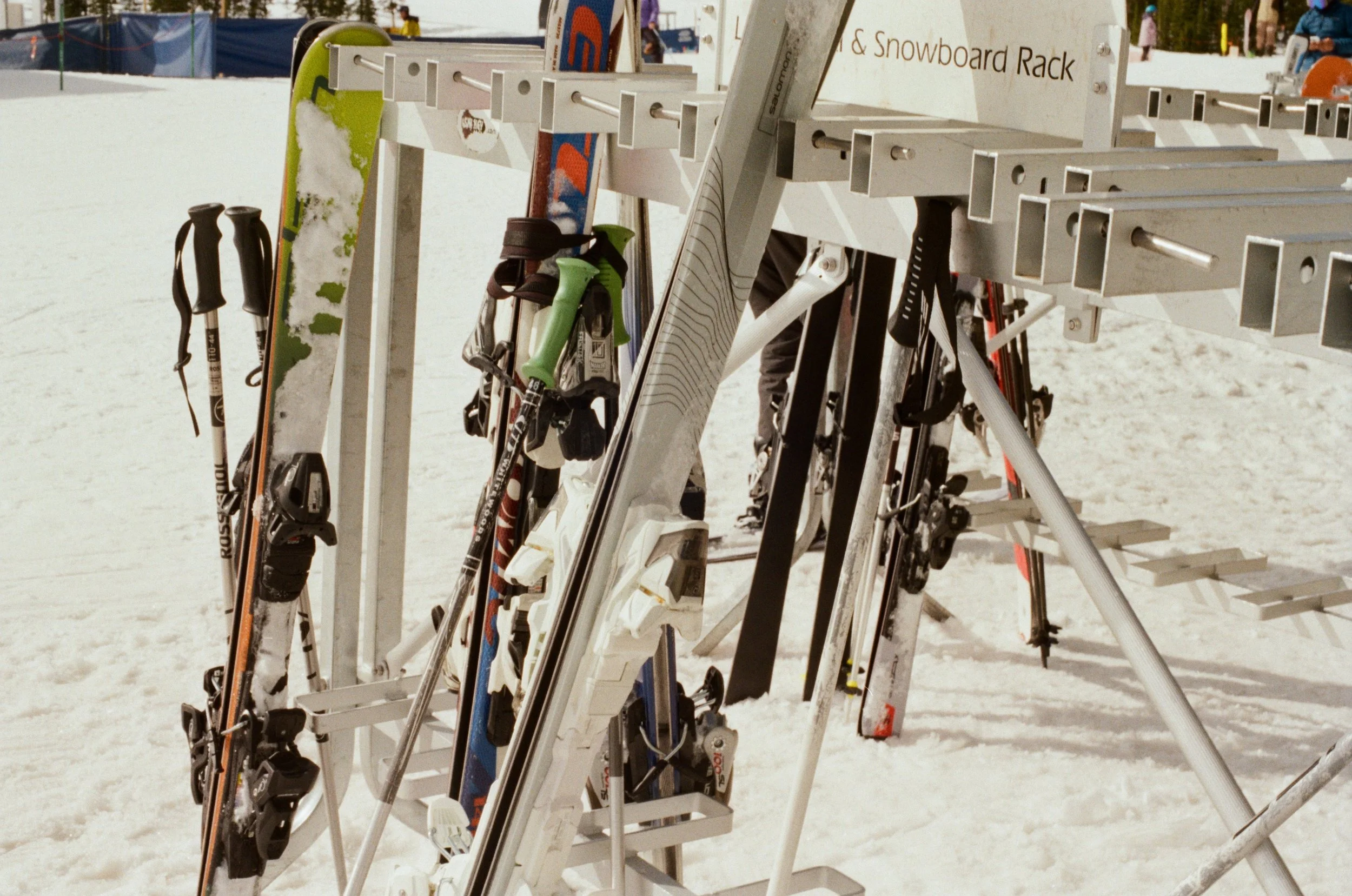 A rack of multiple pairs of skis and ski poles on a snow-covered ground at a ski resort, with people in the background.