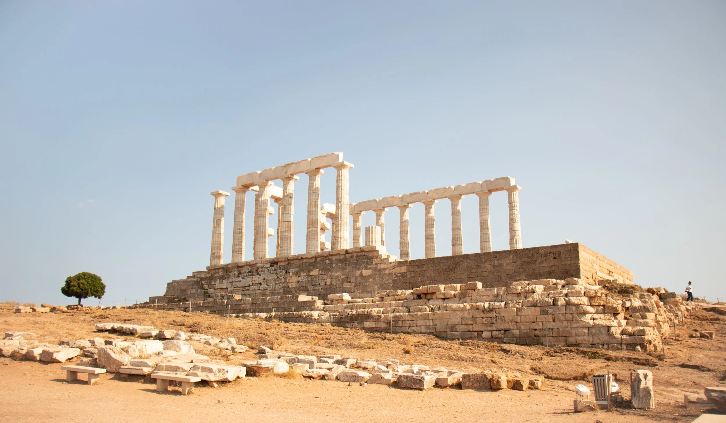 Ancient Greek ruins at the temple of Poseidon with tall white columns on a raised platform, set against a clear blue sky, with a small tree and a person visible on the side.