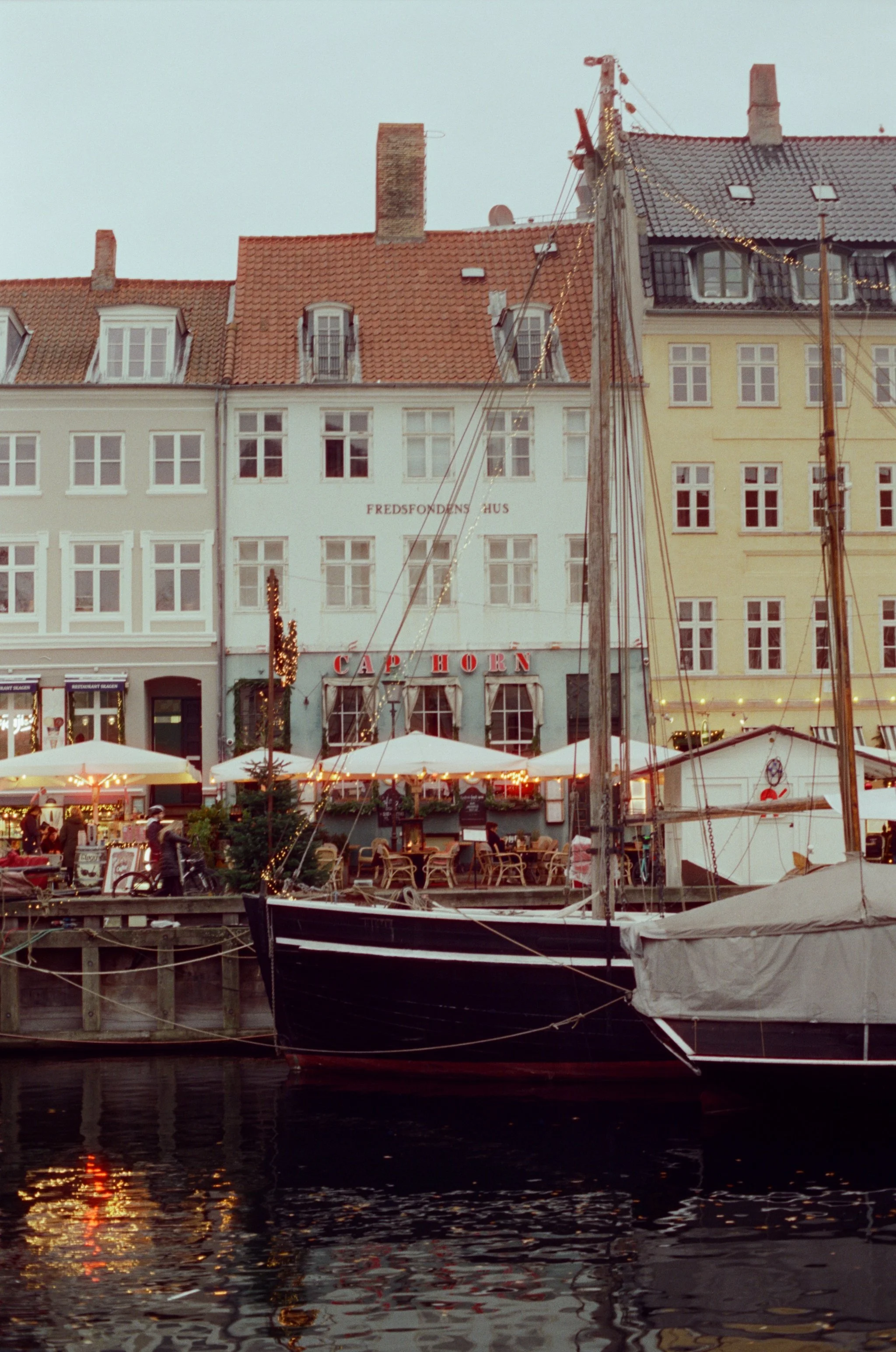 A harbor scene at dusk with boats docked in the water, and buildings with European architecture reflecting in the water. There are outdoor cafes with string lights and people walking by.
