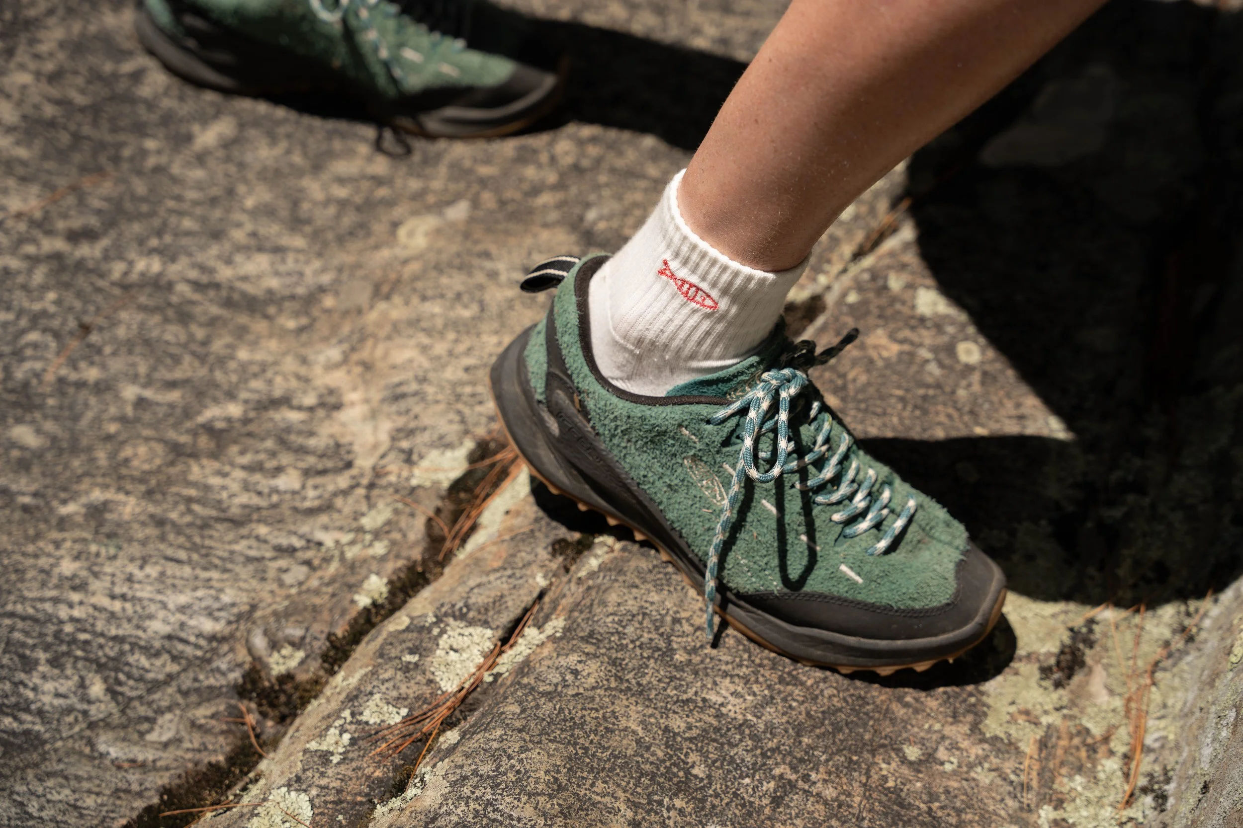 Close-up of a person's foot in a hiking shoe and a white sock with a red fish logo, on a rocky outdoor surface.