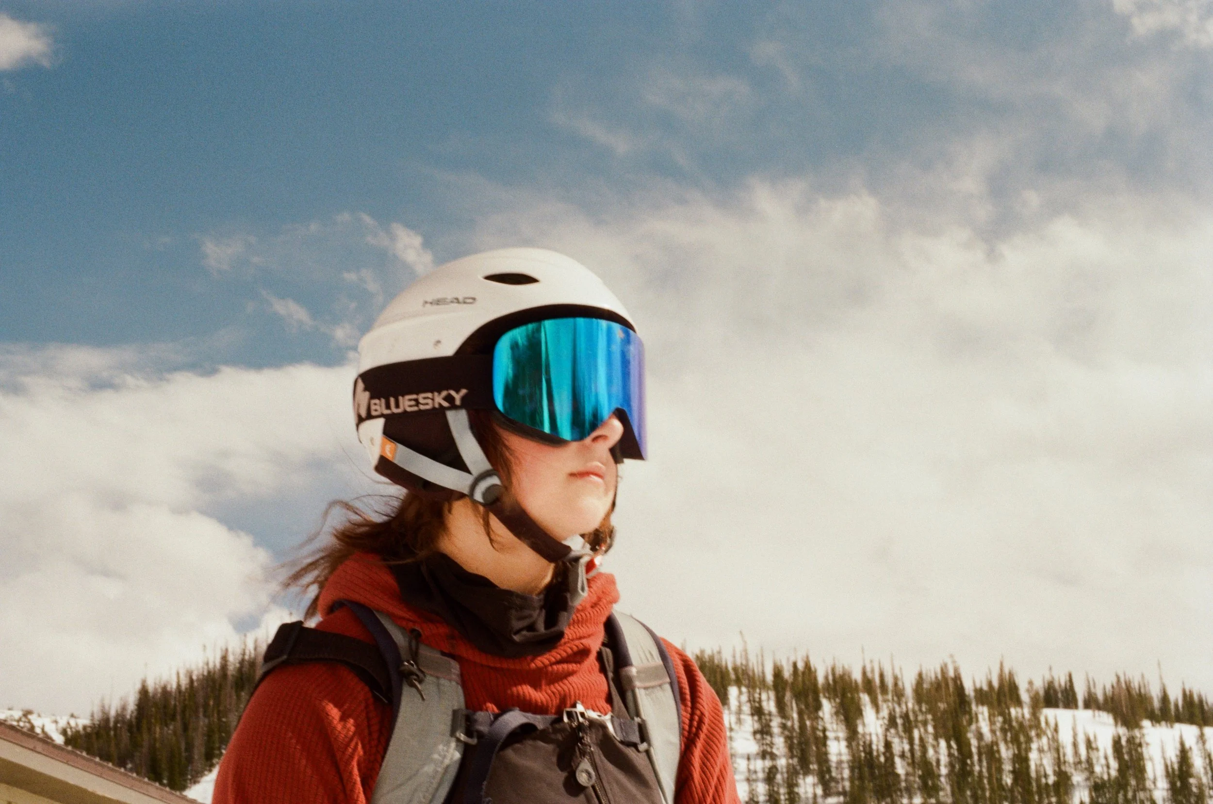 A woman wearing a red jacket, a helmet with a mirrored visor, and a backpack, standing outdoors with snowy landscape and trees in the background under a cloudy sky.