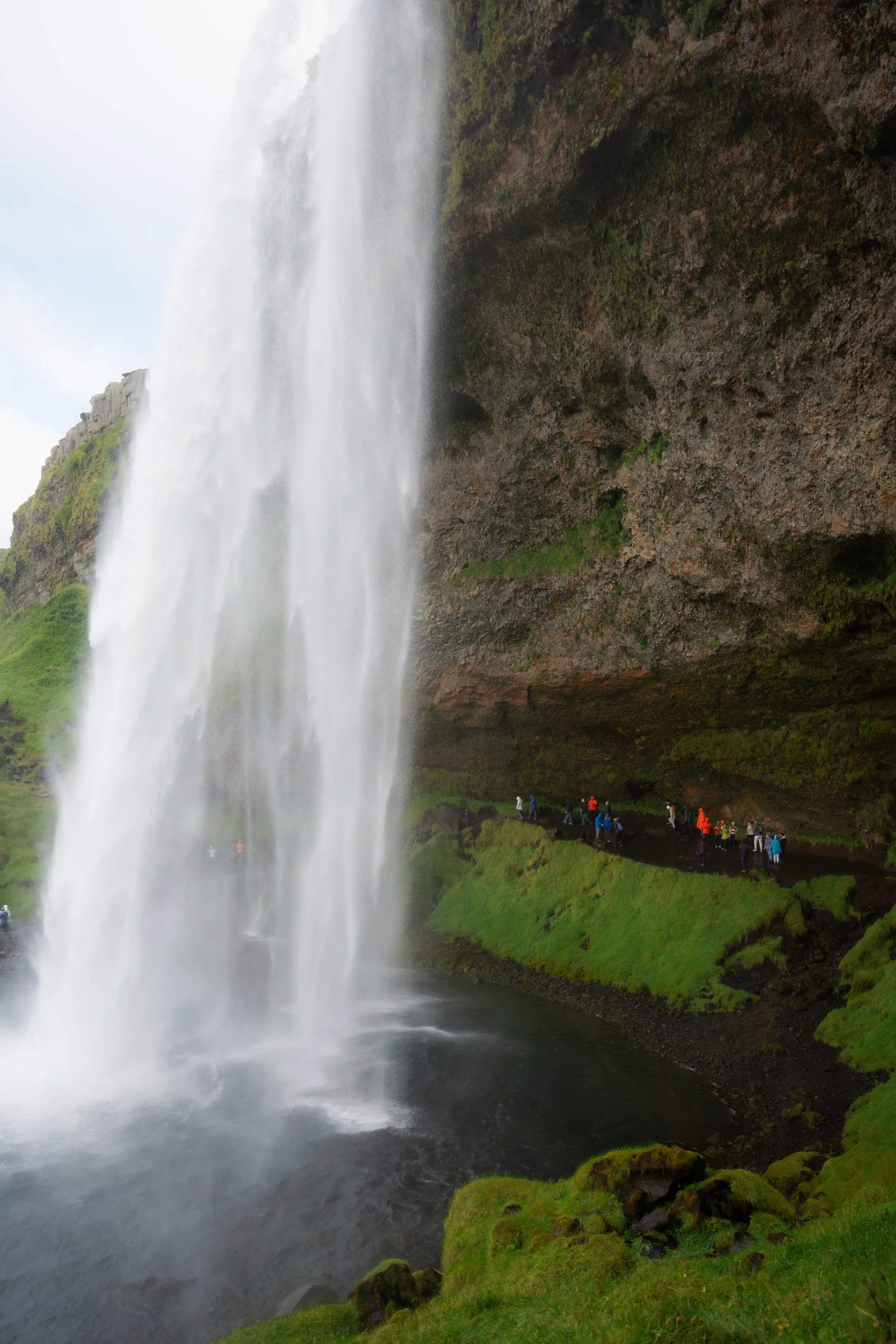 A large waterfall surrounded by greenery, with people standing on a viewing platform at the base of the waterfall.